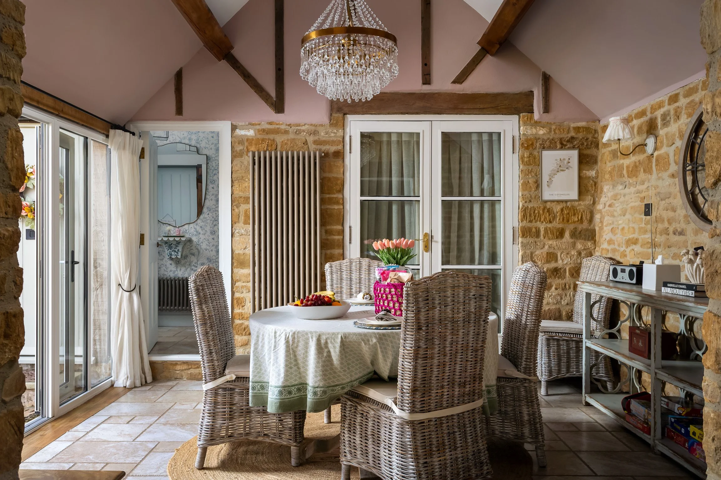 A rustic dining room with a round table covered with a white tablecloth, surrounded by wicker chairs. A vase of pink tulips and a fruit bowl are on the table. The room has exposed brick walls, a chandelier, and a sliding glass door with white curtain
