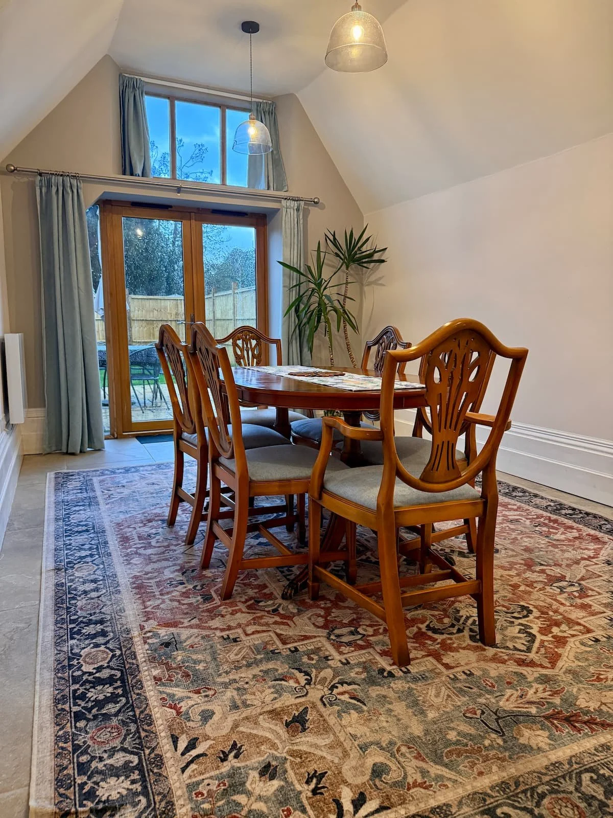 A dining room with a wooden table and six matching chairs, a patterned rug, a large window and glass door, and a potted plant near the wall. The room has sloped ceilings, pendant lights, and blue curtains.
