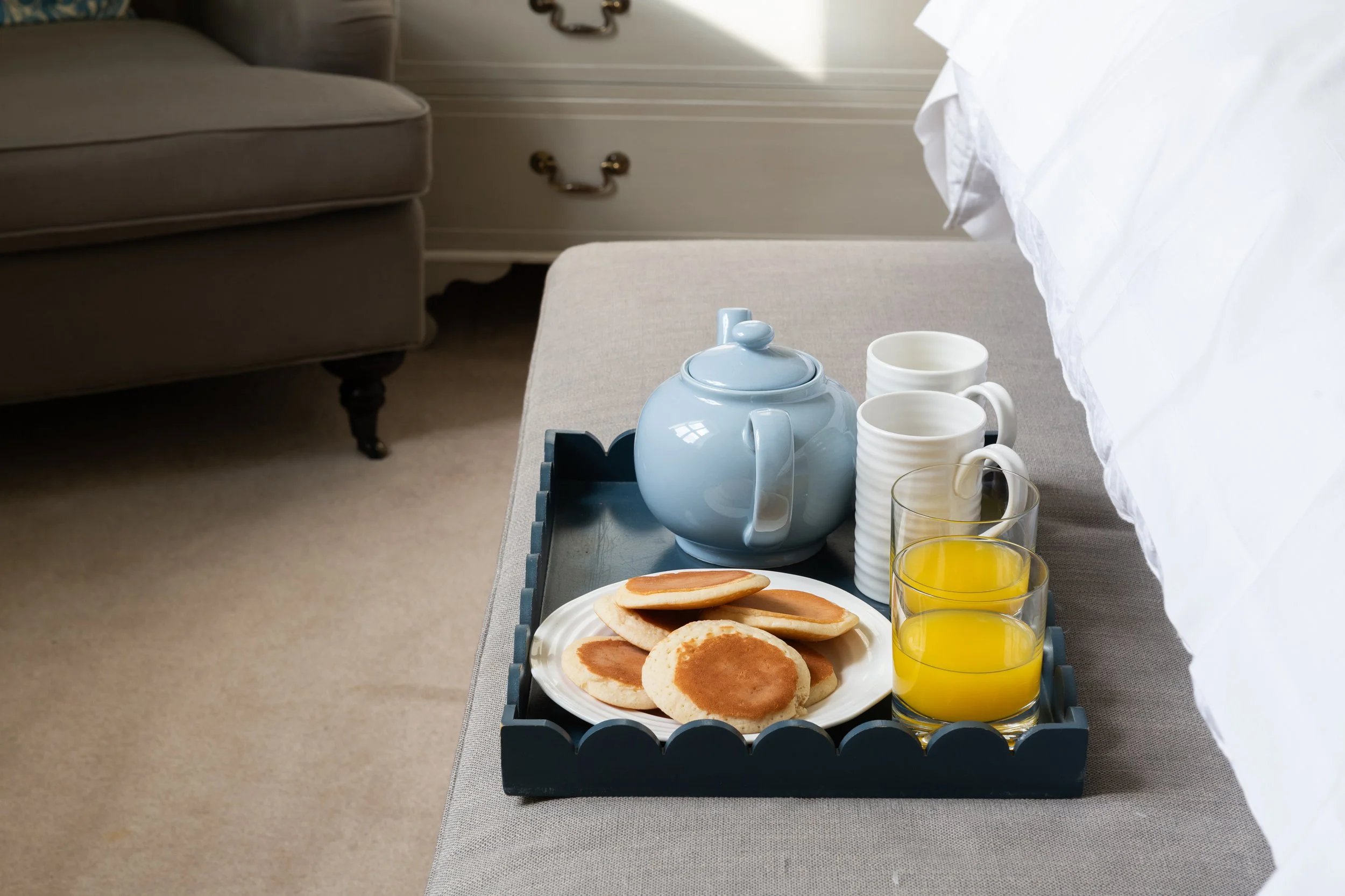 Breakfast tray with a teapot, two white mugs, two glasses of orange juice, and a plate of pancakes, placed on a bed in a bedroom.