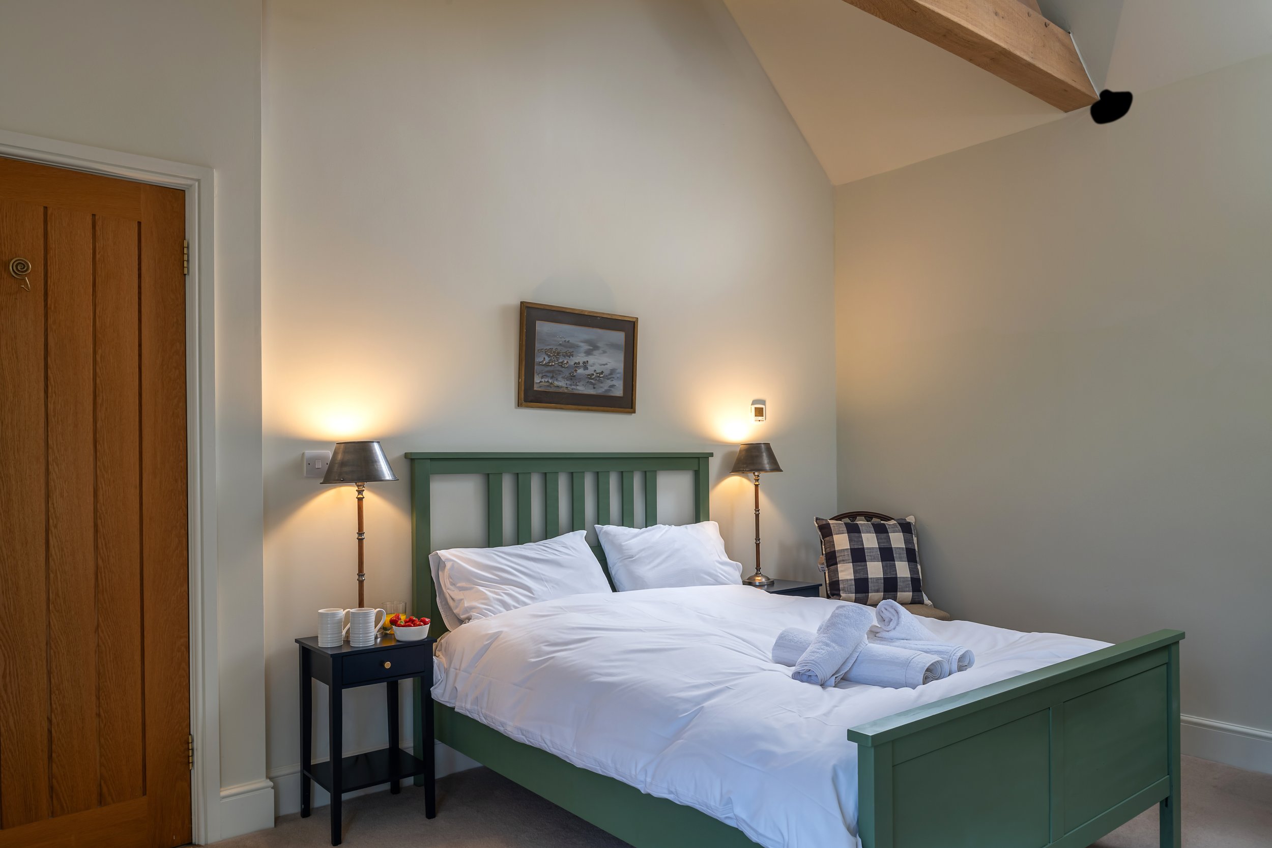 A tidy bedroom with a white bed, green bed frame, and two rolled towels on top. There are two nightstands with lamps, a plaid pillow, and a painting above the bed. Wooden door on the left, beige walls, and a sloped ceiling.
