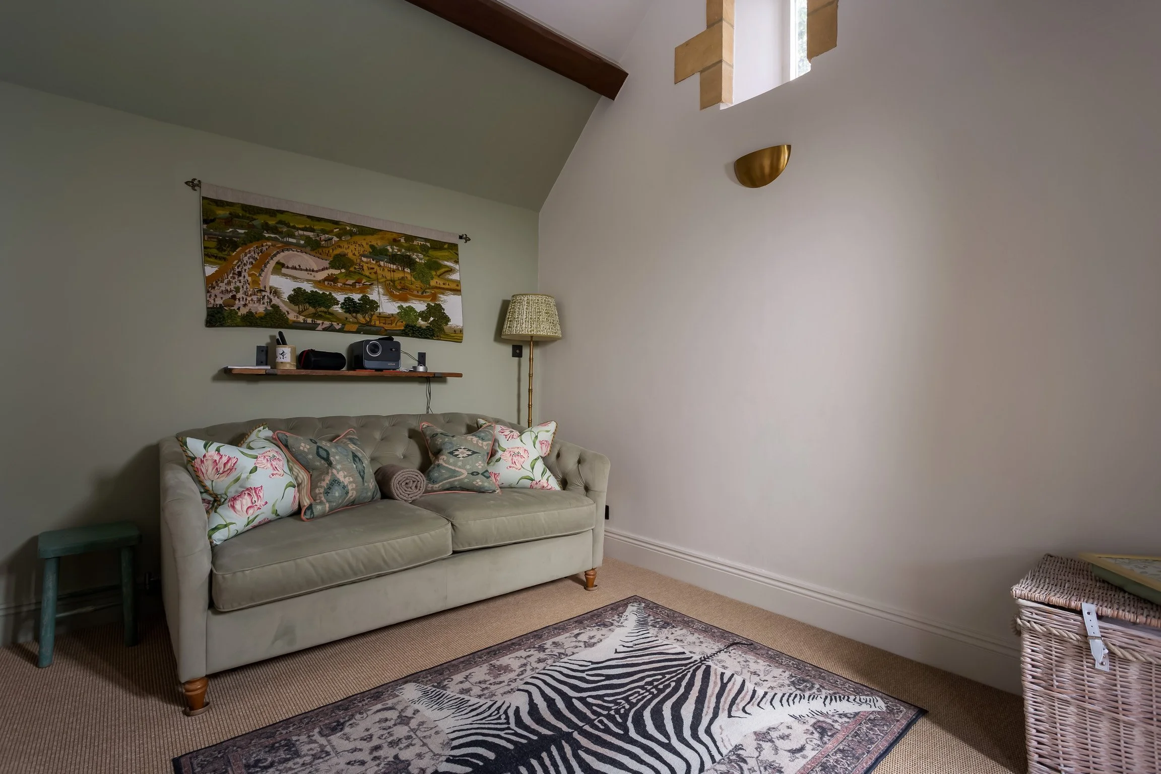 Interior of a living room with a beige tufted sofa adorned with floral and geometric throw pillows, a wooden side table, a vintage standing lamp, a wall-mounted shelf with electronics and a large colorful landscape painting, a patterned rug with zebr