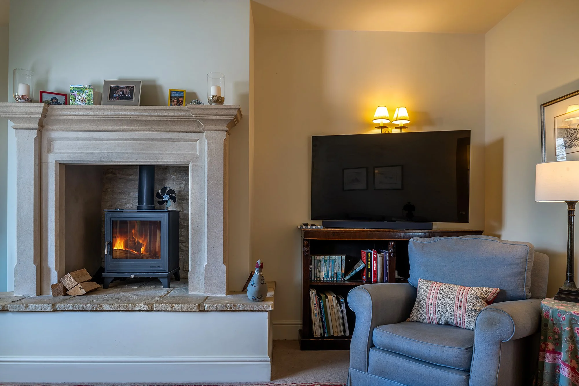 Living room with a fireplace, a large TV on a wooden stand, a beige armchair with a decorative pillow, a table with a floral tablecloth, and wall accents.