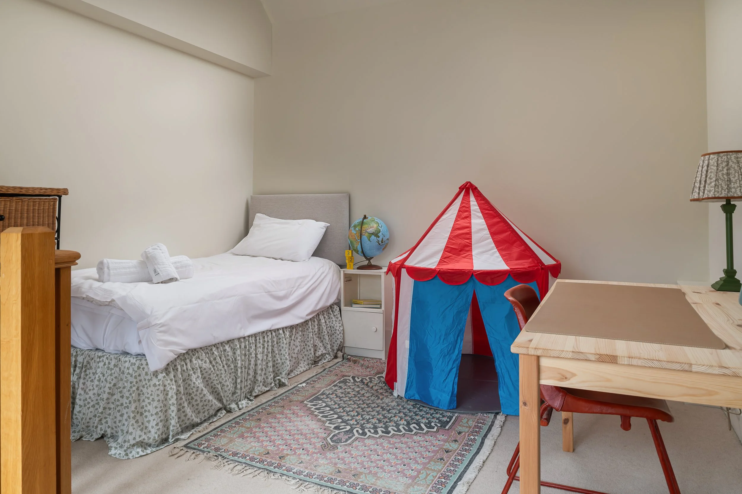 A bedroom with a bed, a small nightstand with a globe, a red and white striped tent, a wooden desk, a brown chair, a lamp, and a colorful rug.