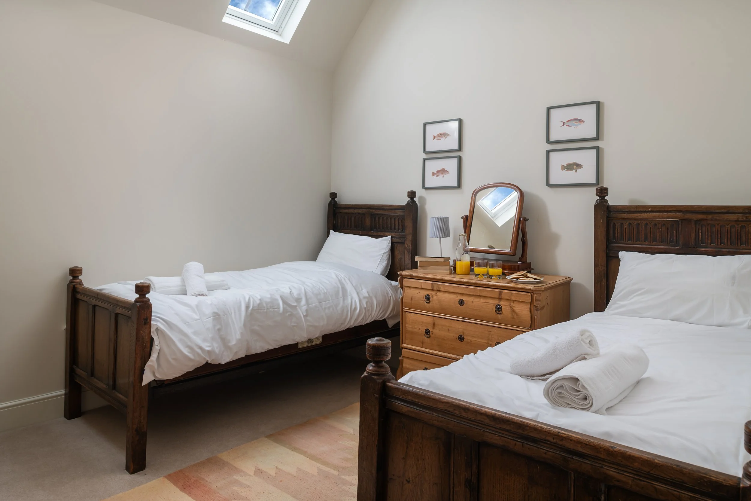 Bedroom with two twin beds with white linens and towels, wooden headboards, a light wood dresser with mirror, framed fish art on the wall, a skylight in the ceiling, and a small table lamp.