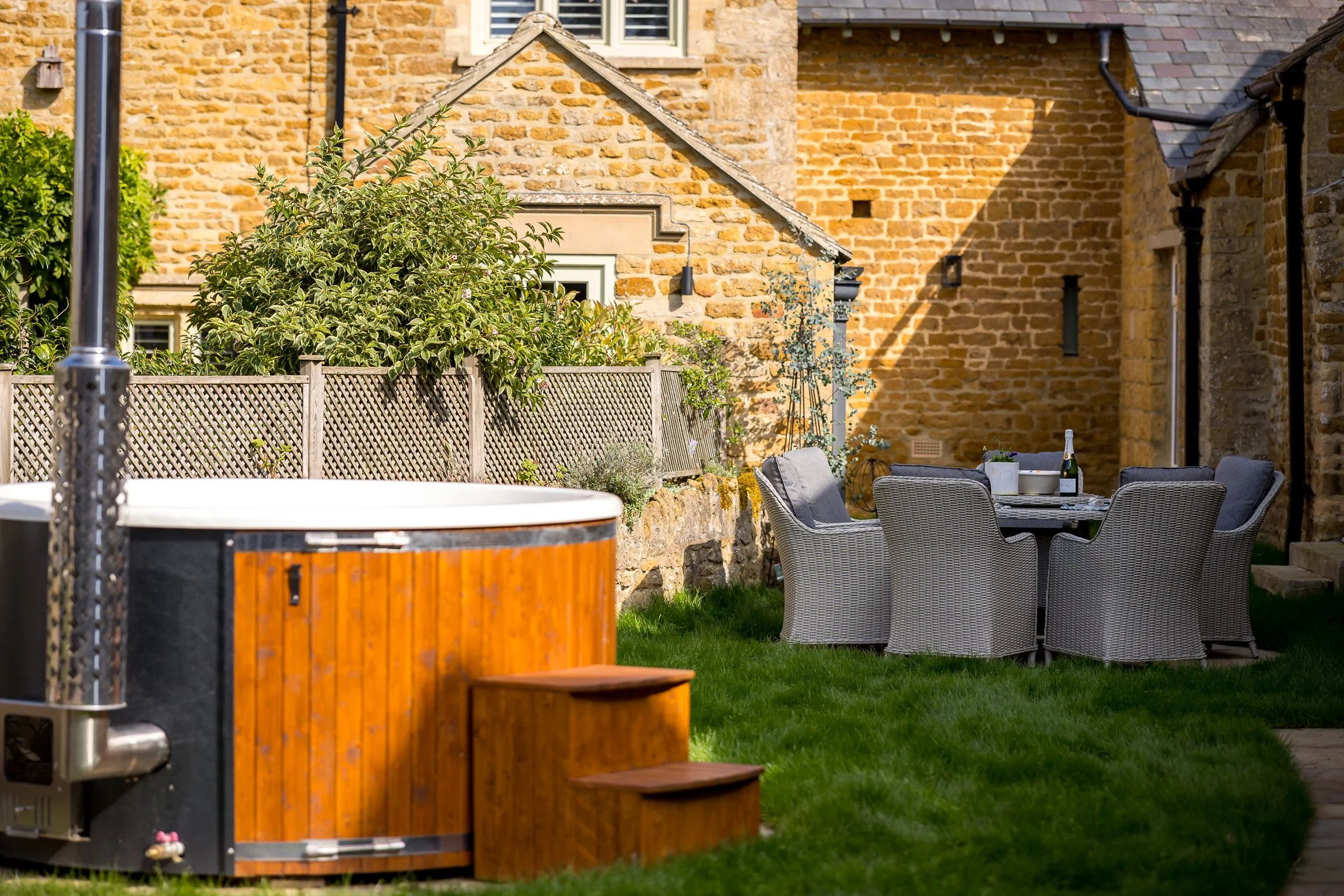 An outdoor backyard scene featuring a hot tub with wooden exterior, a round dining table with six wicker chairs, and a brick house wall with windows and plants.