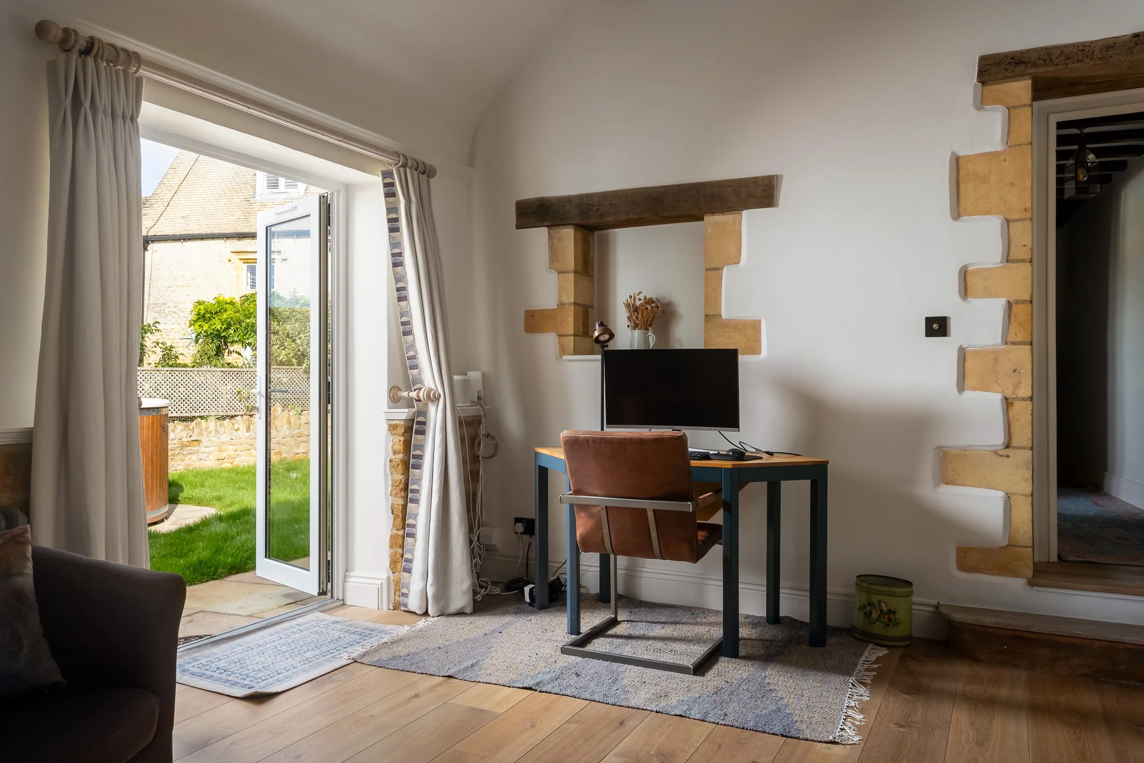 Room with open door to a green garden, small desk with a computer monitor and brown chair, decorative brick wall accents, window with curtains, and hardwood floors.
