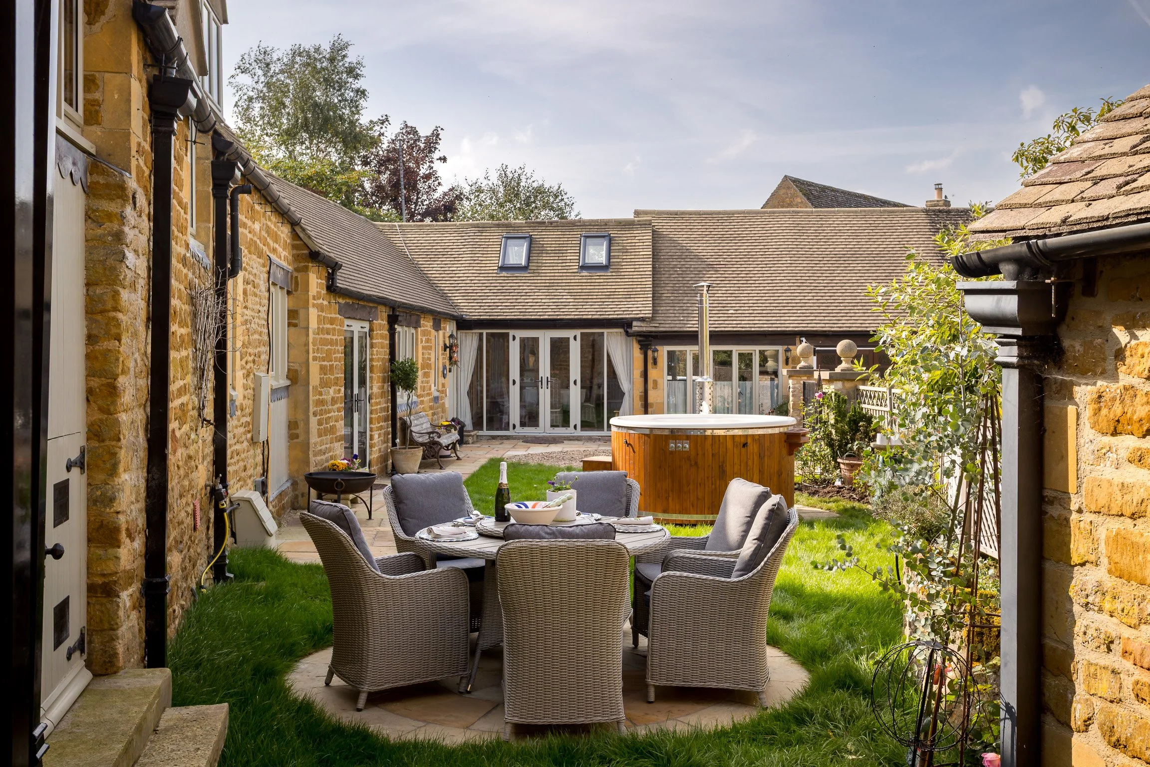 Outdoor patio with a round dining table, six wicker chairs with cushions, and a hot tub in a brick-walled backyard.