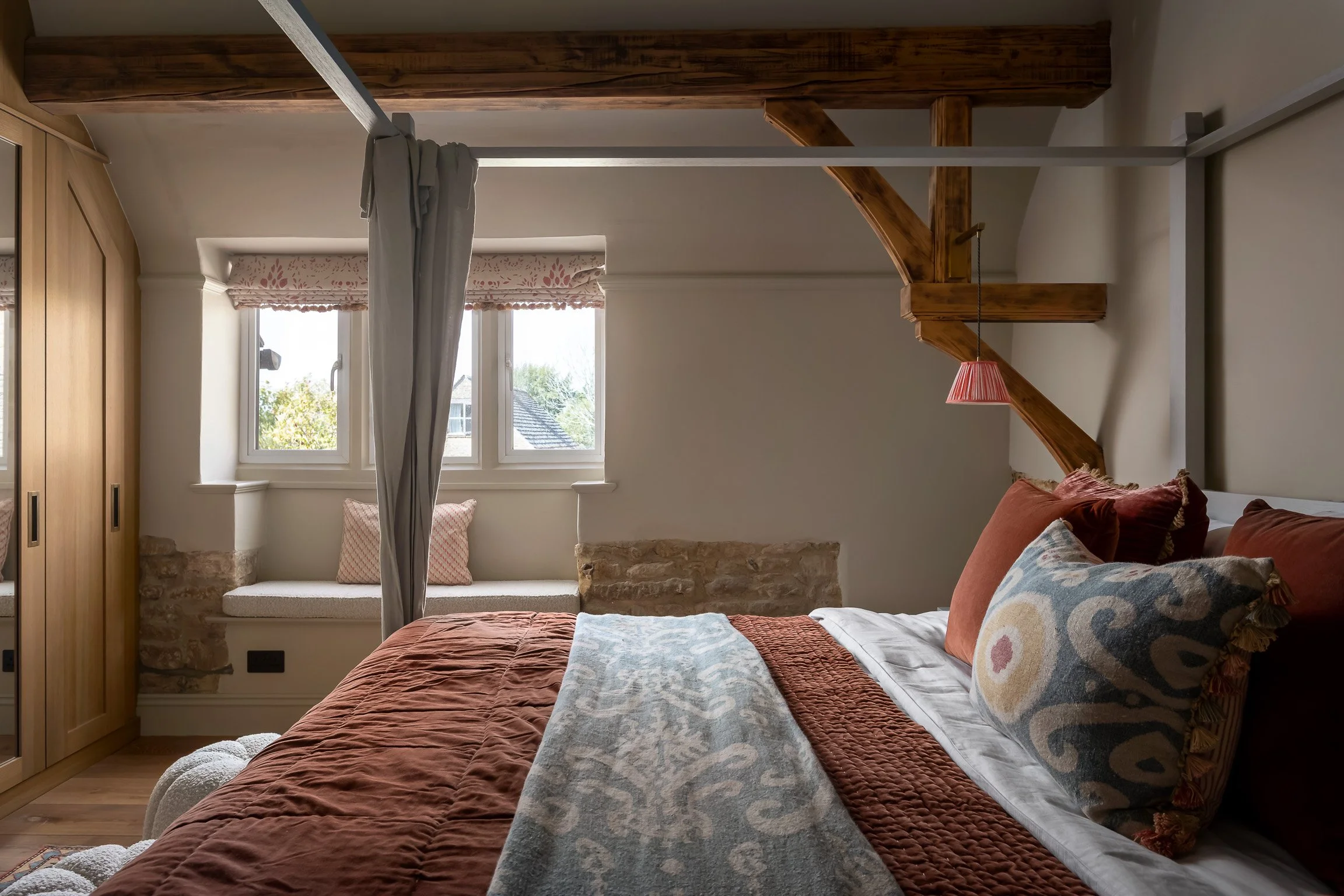 A cozy bedroom with a bed featuring rust-colored bedding and decorative pillows, a window seat with a cushion and pillow, a large window with a floral valance, wooden beams, and a hanging pink lamp.