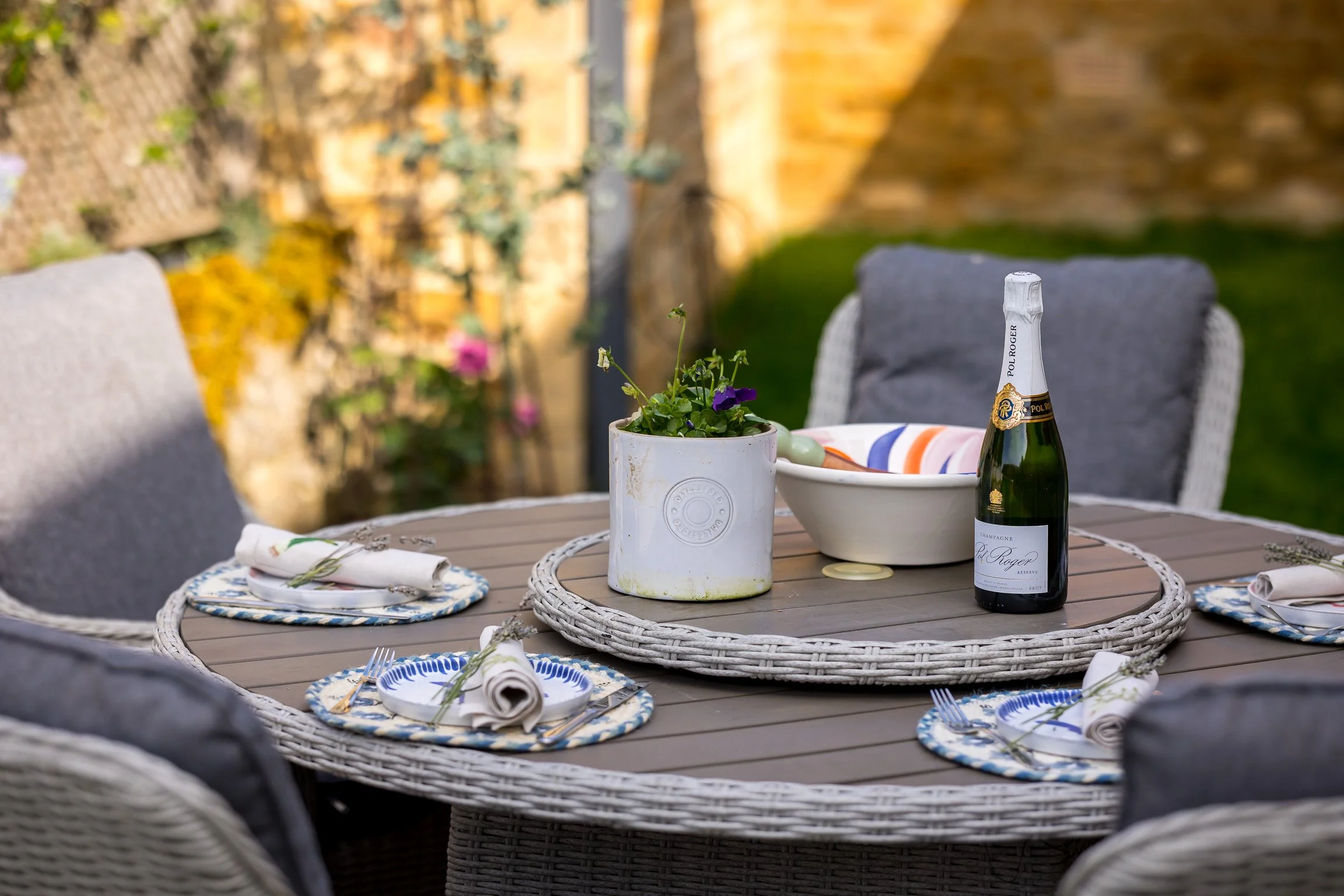 Outdoor patio table set for a meal with plates, napkins, and utensils, a bottle of champagne, a bowl, and a potted plant with purple flowers, with cushioned wicker chairs and a garden background.
