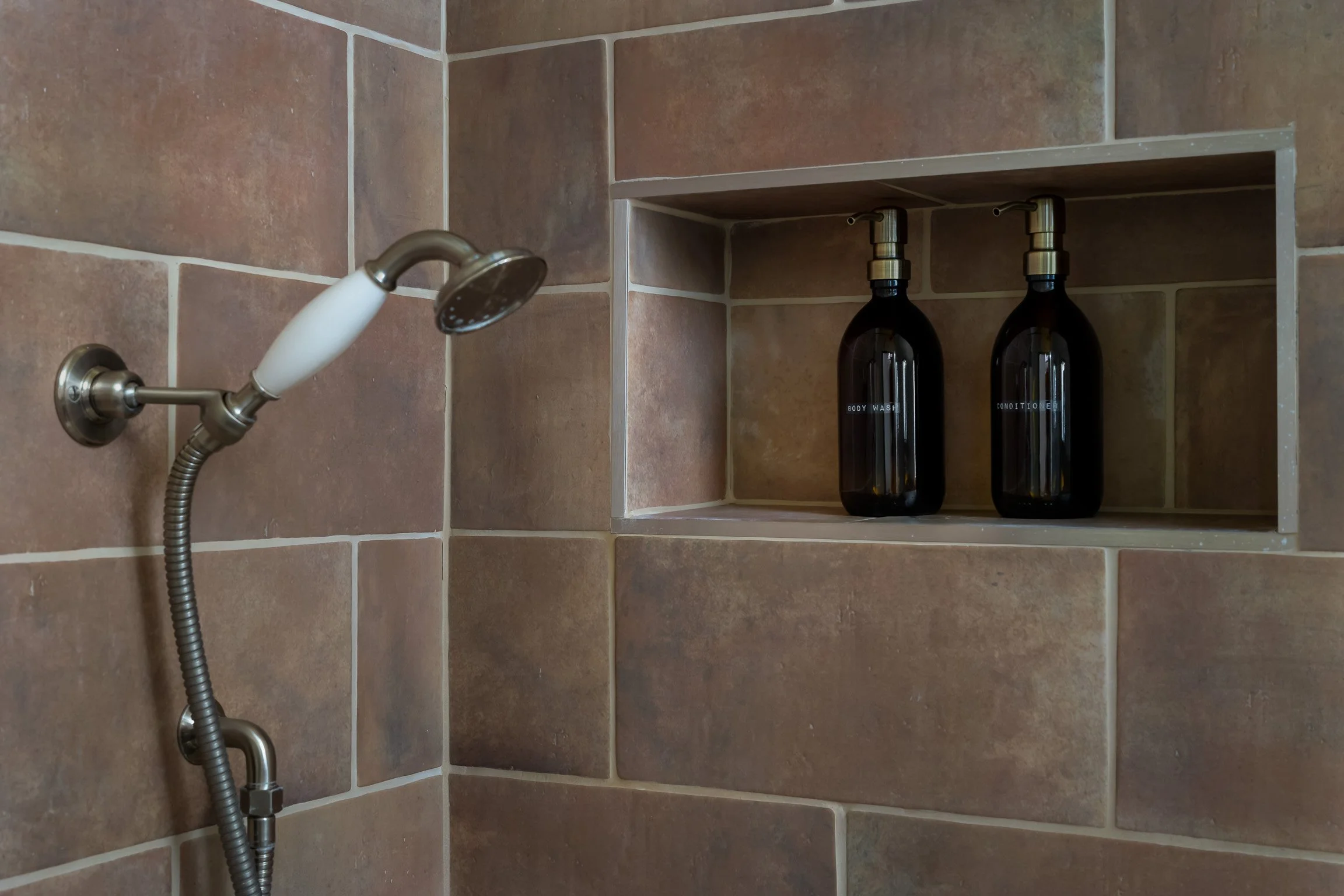 Close-up of a shower corner with brown tiled wall, a shower head with a white handle, and a built-in shelf holding two dark bottles labeled 'Body Wash' and 'Conditioner'.