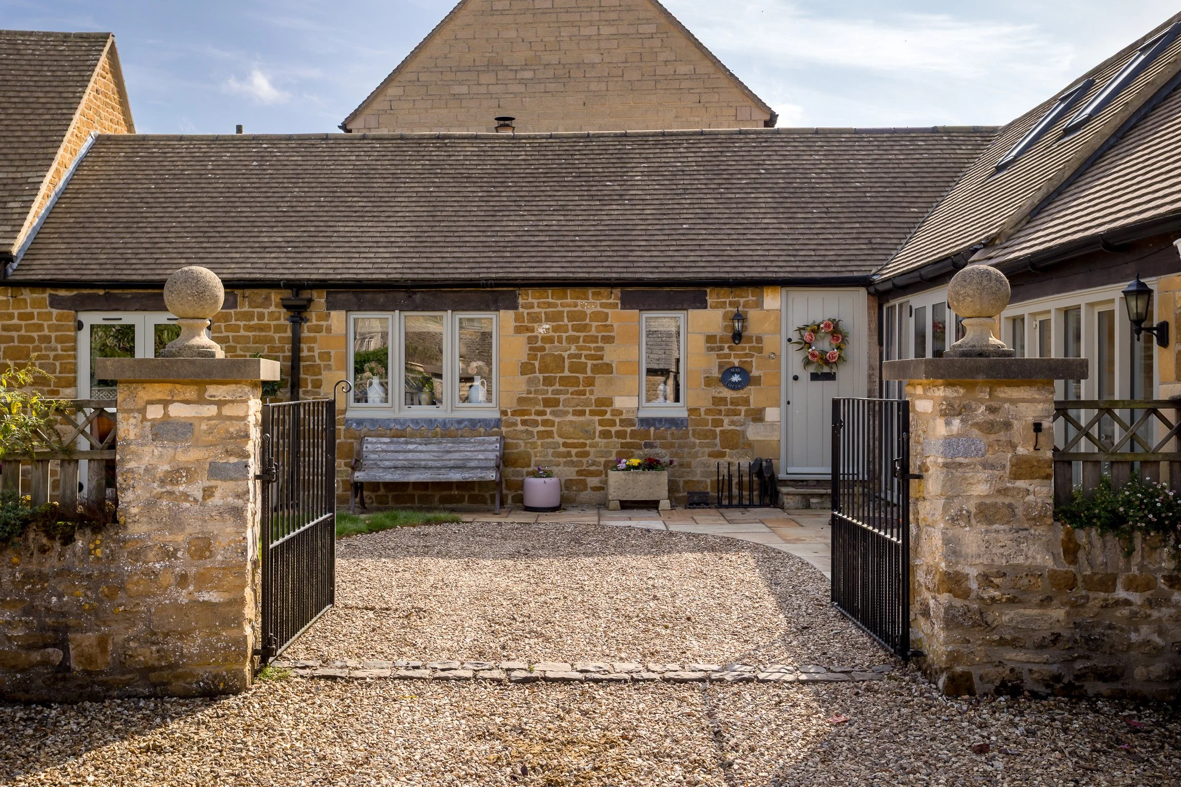 Front view of a stone house with a gravel driveway and black iron gate, decorated with a flower wreath on the door, benches, flower pots, and outdoor lighting.