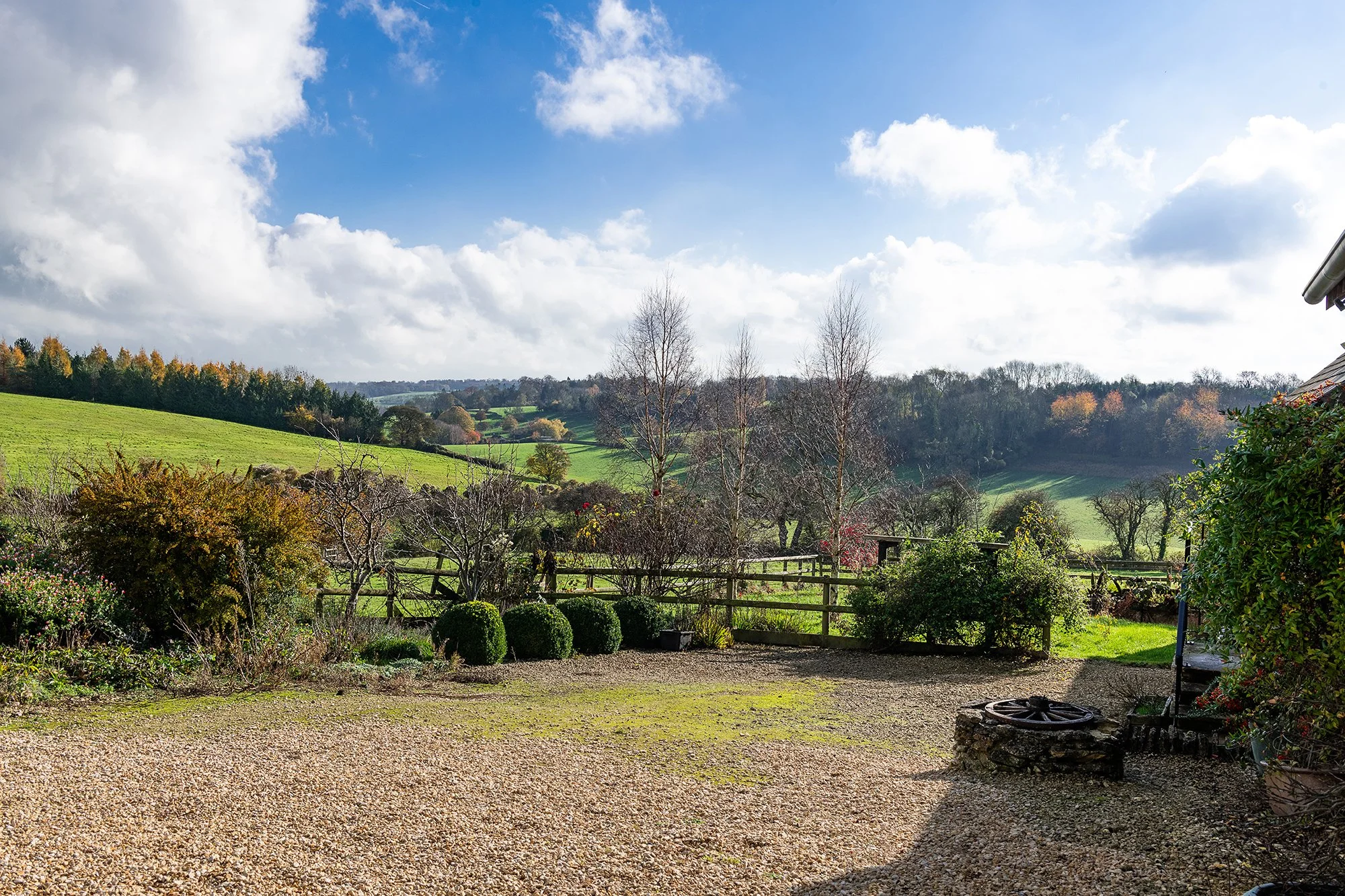 Scenic view of a rural landscape with rolling green hills, trees, and partly cloudy blue sky, as seen from a gravel patio area with bushes and a wooden fence in the foreground.
