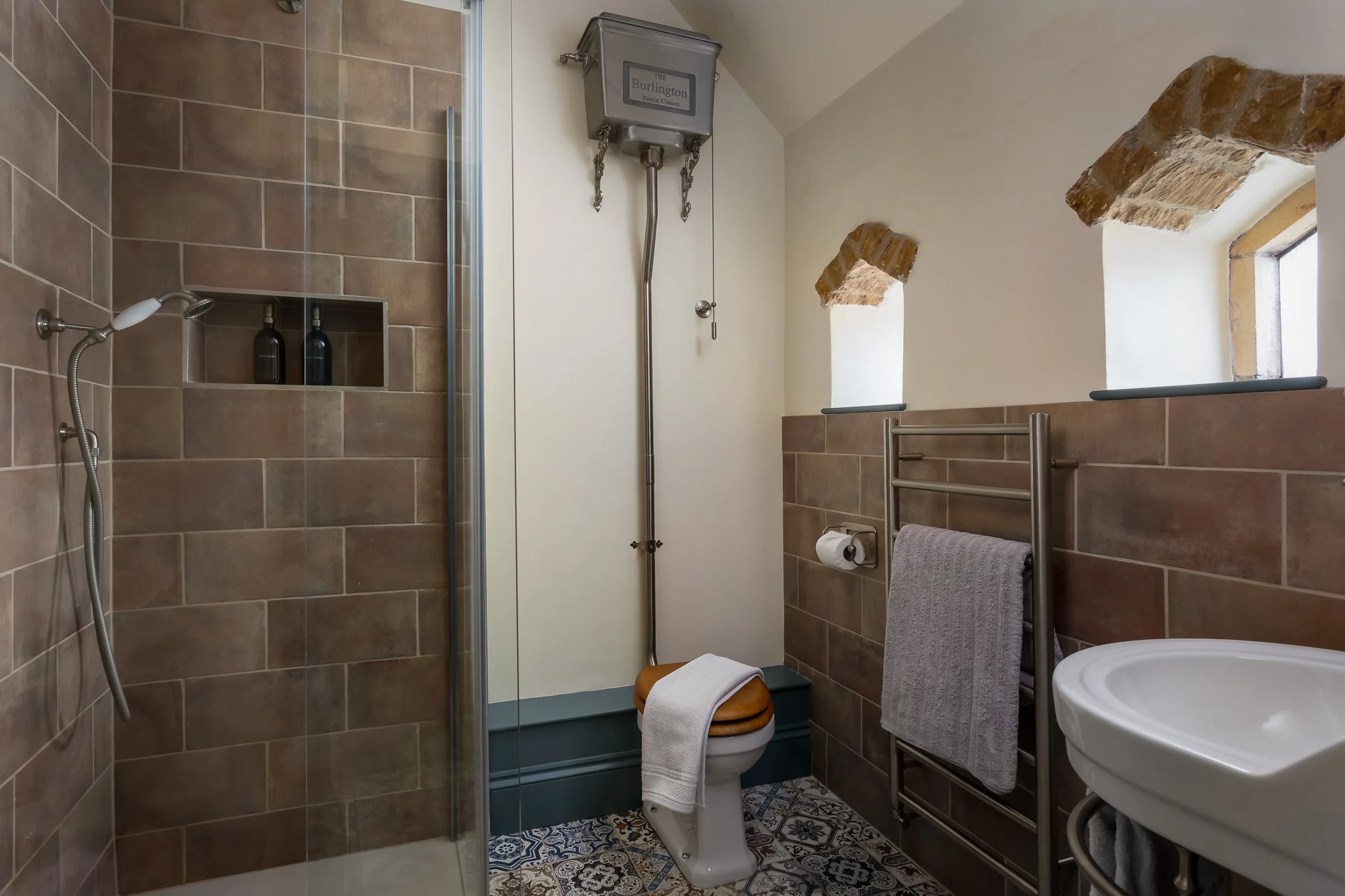 Bathroom with brown tiled walls, a walk-in shower with glass door, a toilet with a wooden seat and towel draped over the lid, a small metal towel rack with a gray towel, two small windows with brick arches, and a white sink.