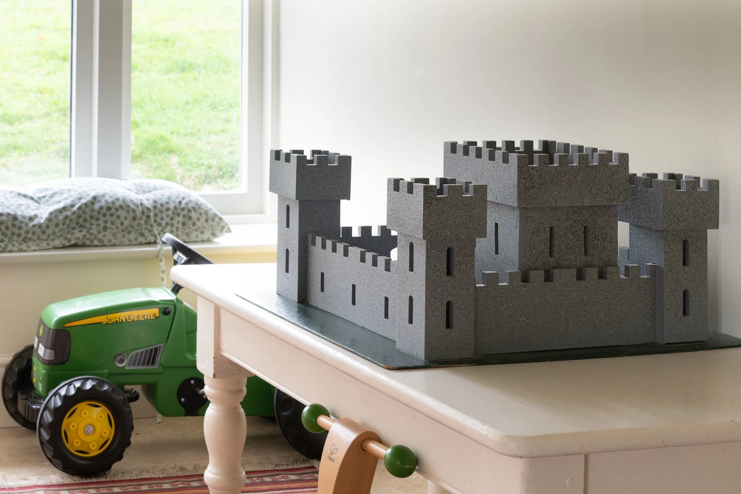 Children's play area with a gray foam castle on a white table, a green toy tractor, a window with green grass outside, a pillow with a polka dot pillowcase, and part of a red-striped rug.