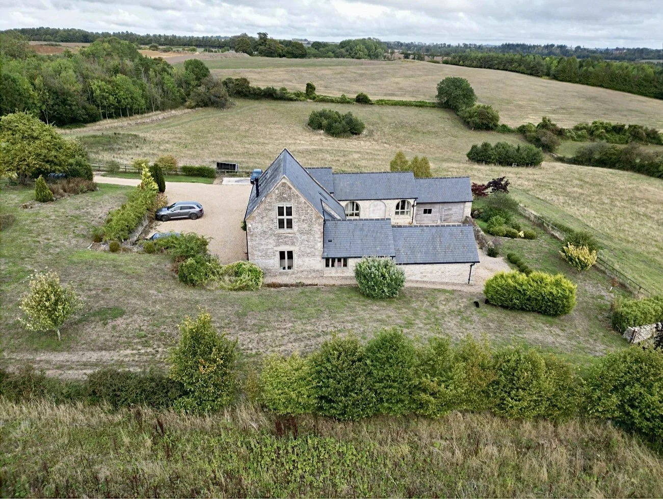 An aerial view of a stone house with a slate roof, surrounded by a gravel driveway and lush green landscape, including trees and open fields, under a cloudy sky.
