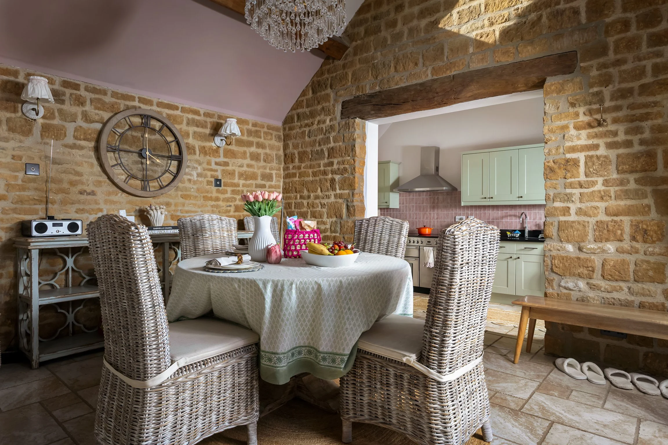 A dining room with a round table covered in a lace tablecloth, surrounded by wicker chairs. The table has a white vase with pink tulips, a bowl of fruit, and some small decorations. The walls are exposed brick, with wall-mounted lamps, a large wall c
