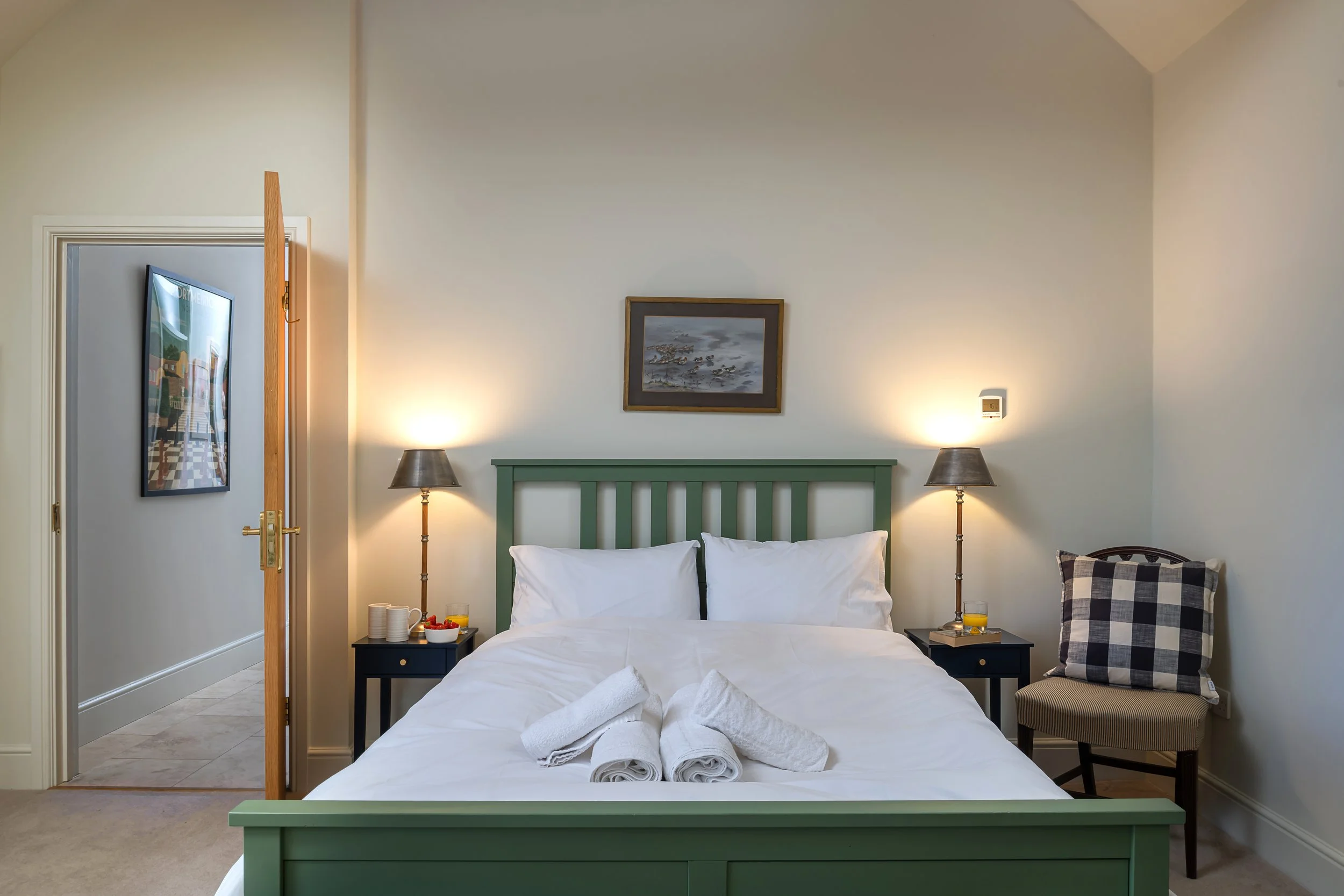 A neatly made bed with white linens and towels, green headboard, two pillows, in a well-lit bedroom with two bedside lamps, a chair with cushions, and wall art.