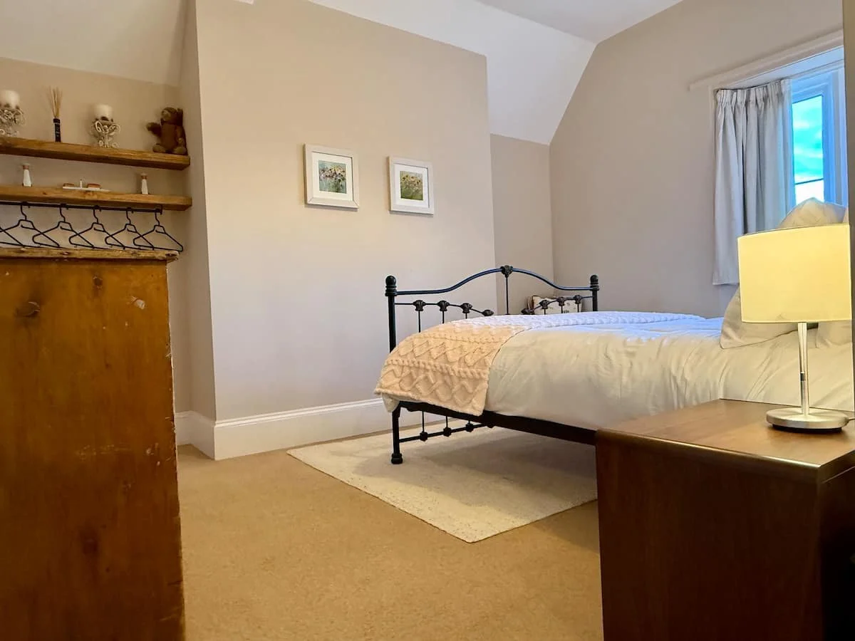 A cozy bedroom with a white bed, beige bedding, a wooden nightstand with a lamp, a white rug, and a window with white curtains, walls decorated with small framed pictures, and a shelf with decorative items.