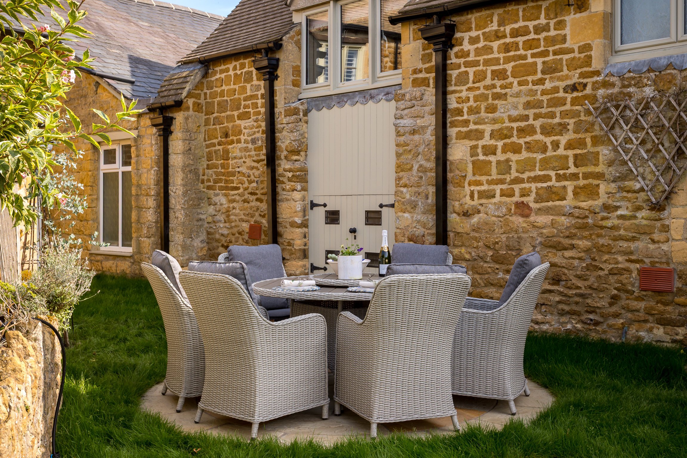 Outdoor patio area with a round table surrounded by eight wicker chairs, two of which have gray cushions, on a grassy lawn. The table has a white flower pot, a bottle of wine, and some plates. Behind it is a brick building with windows, a closed whit