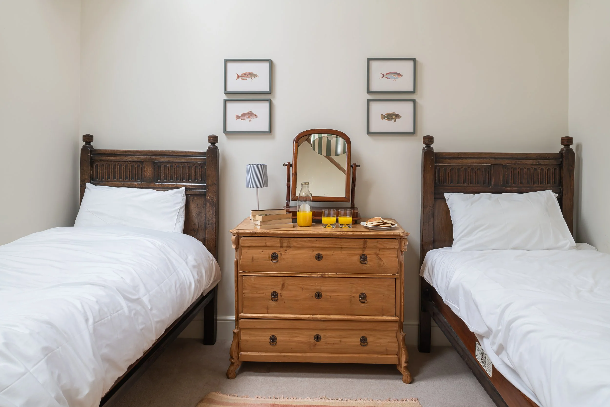 A cozy bedroom featuring two wooden twin beds with white bedding, a wooden dresser with a mirror in the center, and framed fish artwork on the wall above each bed. The dresser has a small lamp, a stack of books, a glass pitcher of orange juice, two g