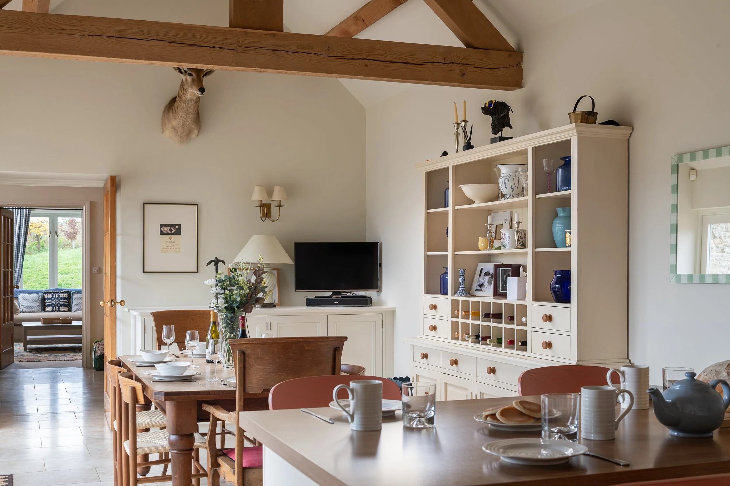 Interior of a dining room with a wooden dining table set with dishes, cups, and glasses. In the background, a white cabinet with decorative blue and white items and a small TV. A faux deer head is mounted high on the wall, and there is a window showi