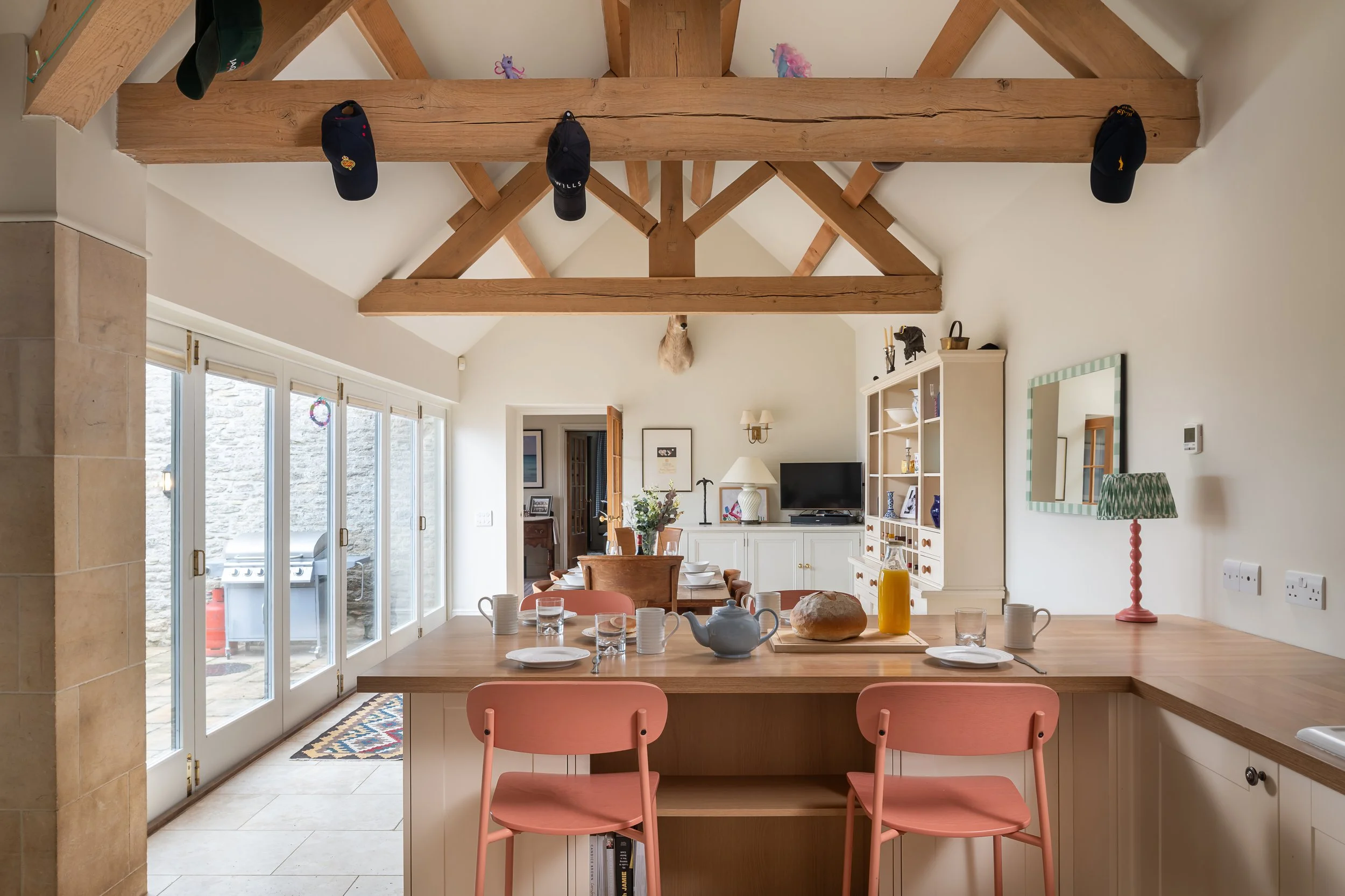 Dining area with a wooden table set with bowls, cups, and a teapot, surrounded by pink chairs. Large glass sliding doors lead to a patio with a grill outside. Ceiling with exposed wooden beams and hats hanging from them. In the background, a white ca
