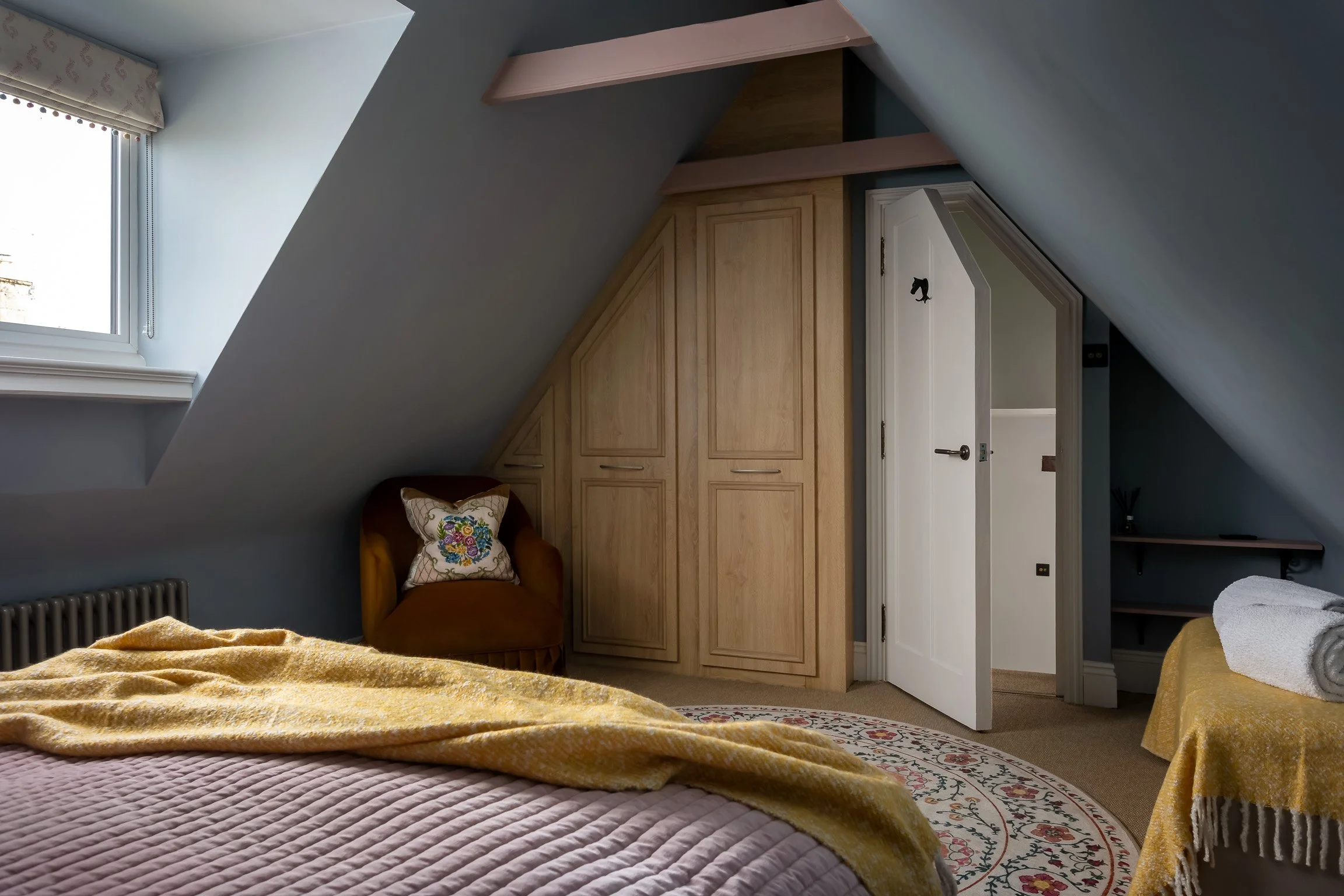 A bedroom with an angled ceiling, a window with a blinds and valance, a bed with a pink quilt and a yellow blanket, a round floral rug, a yellow upholstered chair with a embroidered pillow, a wooden wardrobe with angled doors, and a small white door 