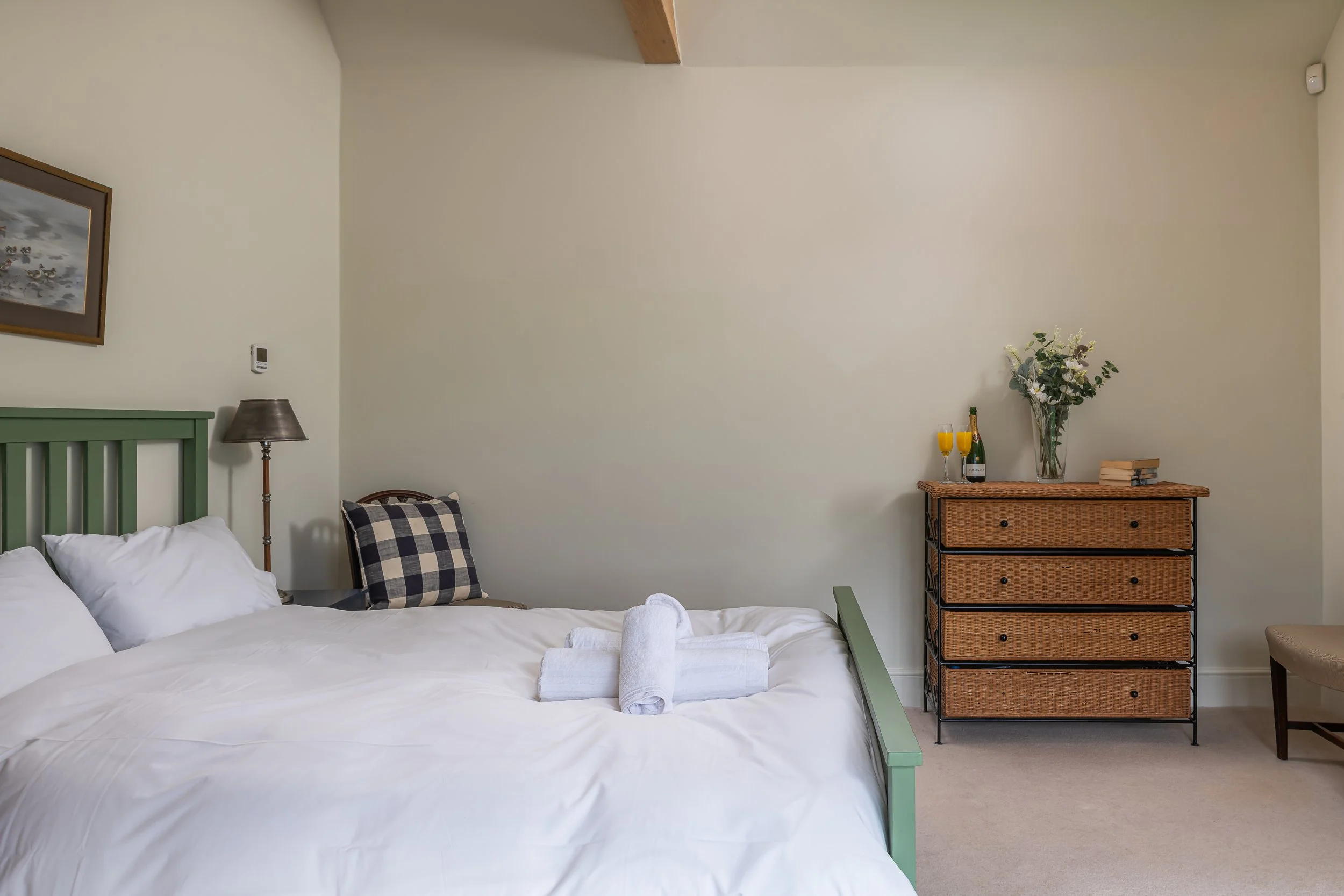 A neatly made bed with white sheets, pillows, and towels in a bedroom. A wooden dresser with a vase of flowers, two glasses of orange juice, a bottle of wine, and some books is beside the bed. There's also a chair on the right side of the dresser.