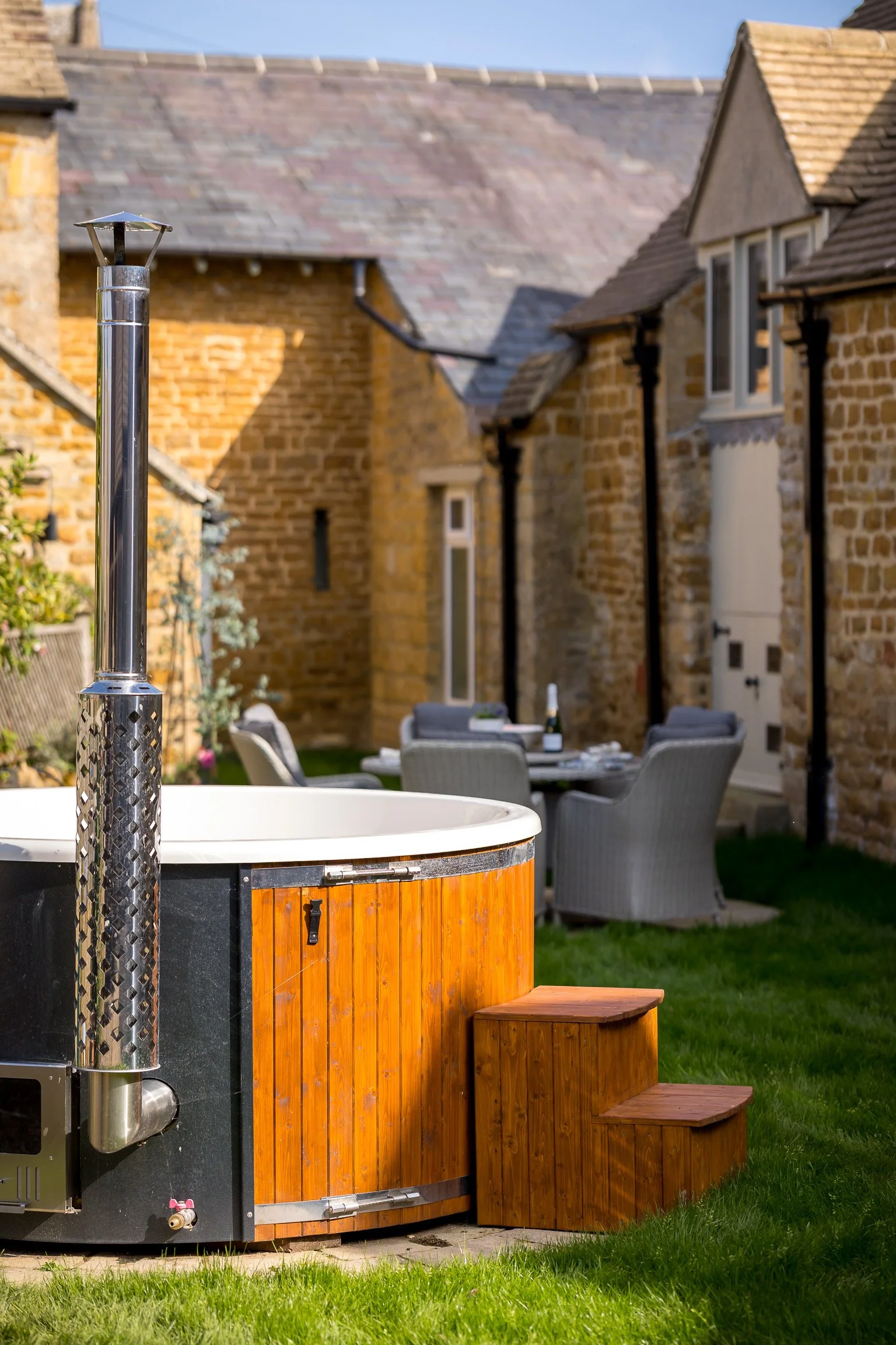 An outdoor backyard scene with a hot tub, wooden steps, and a dining table with chairs, set against brick houses under a clear sky.