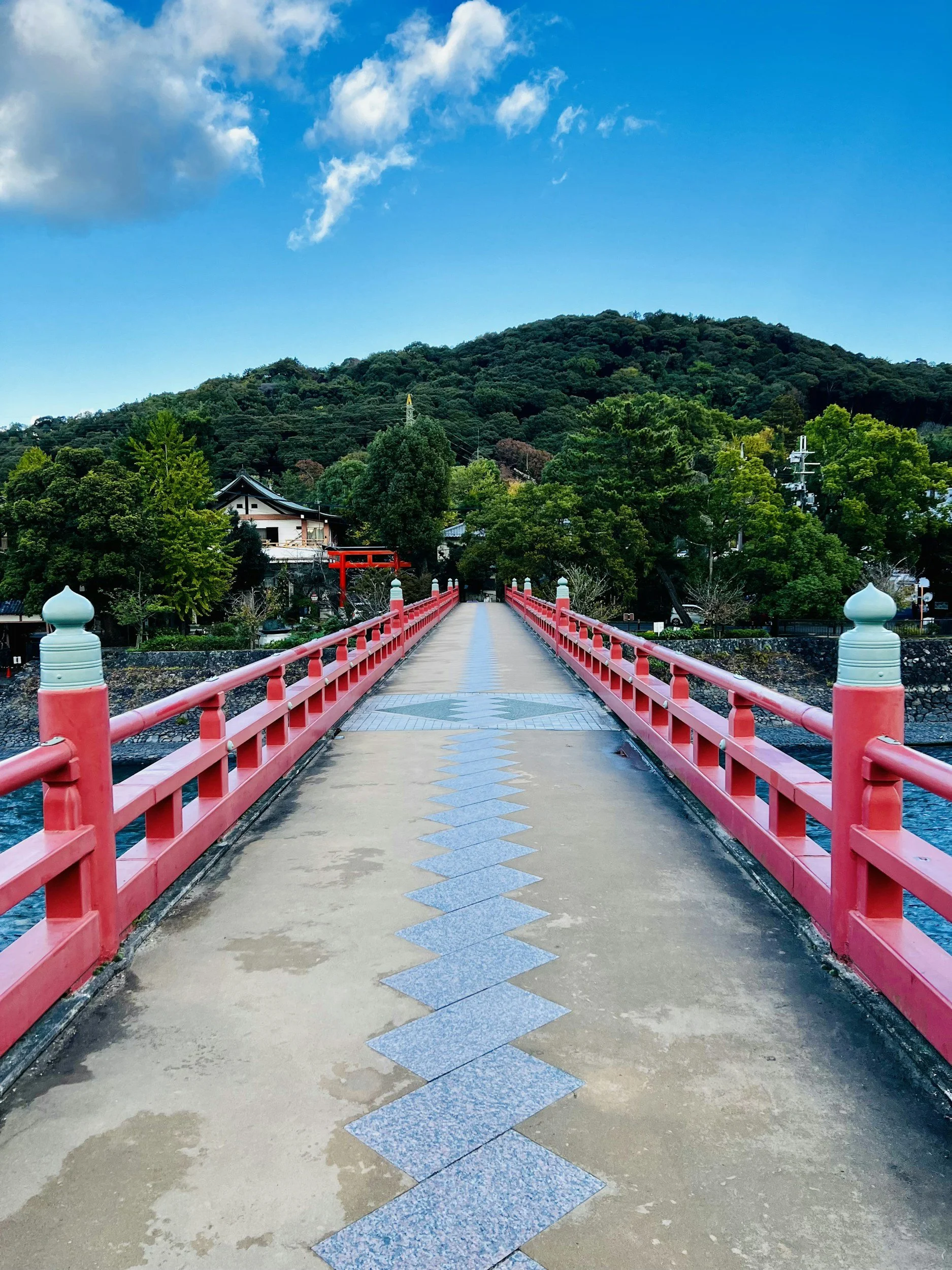 A red bridge crossing over water with a mountain and lush green trees in the background under a partly cloudy blue sky.
