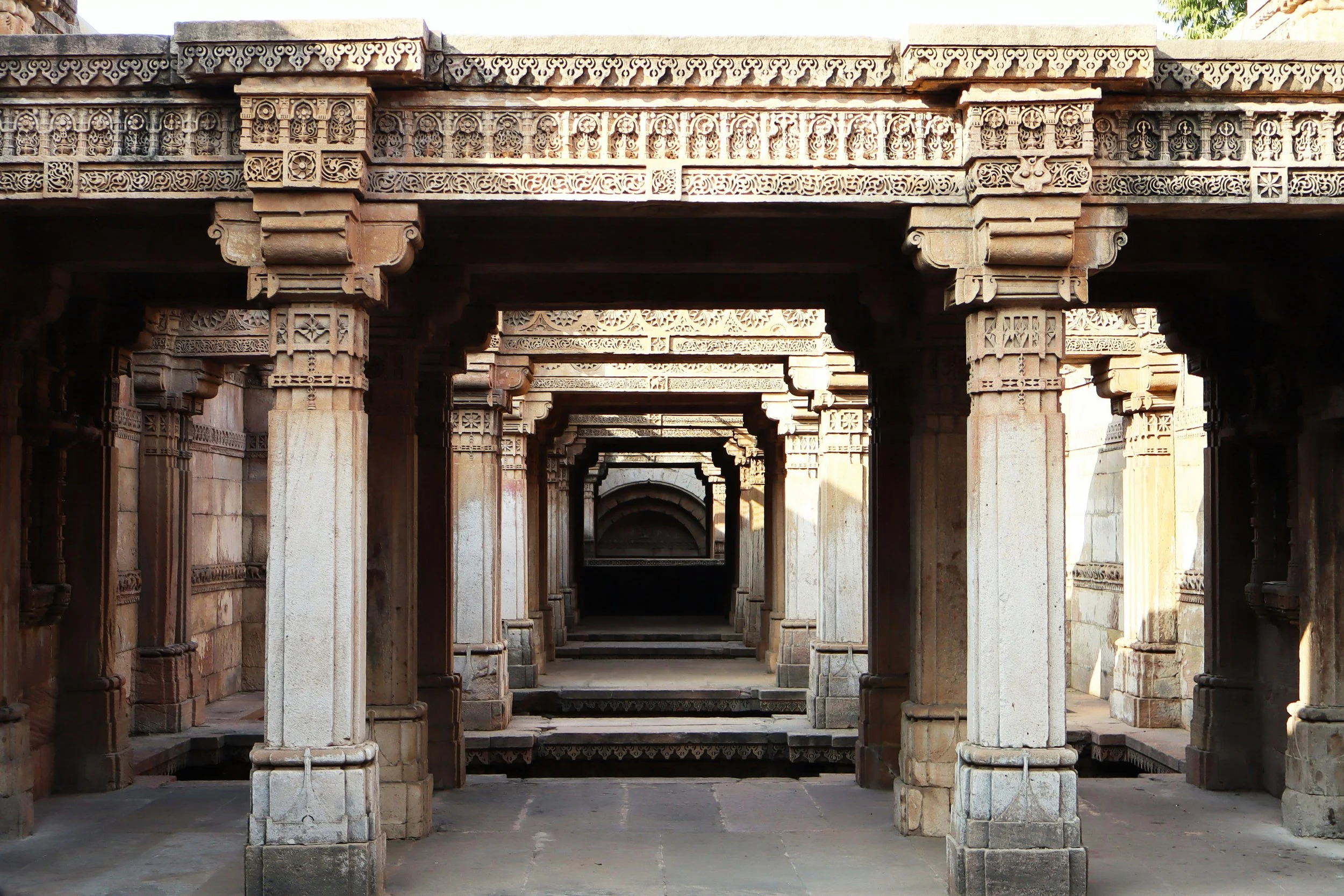 Ancient stone architectural corridor with multiple carved pillars and detailed decorative friezes, creating a repetitive pattern.