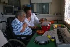 A caregiver and an elderly woman in a striped shirt sitting at a table, engaged in an activity together with small cups and objects, in a room with a window, keyboard, and turntable.