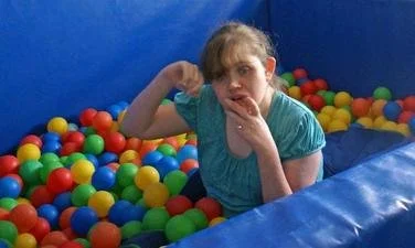 Girl sitting in a ball pit with colorful plastic balls, blue padded walls surrounding her.