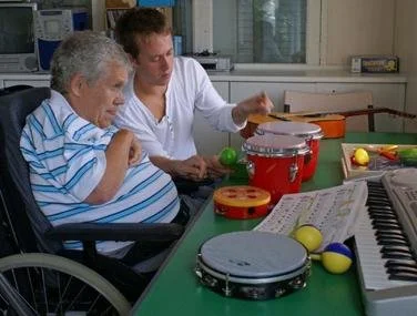 A young man in a white shirt assisting an elderly woman in a blue and white striped shirt at a table with musical instruments and toys.