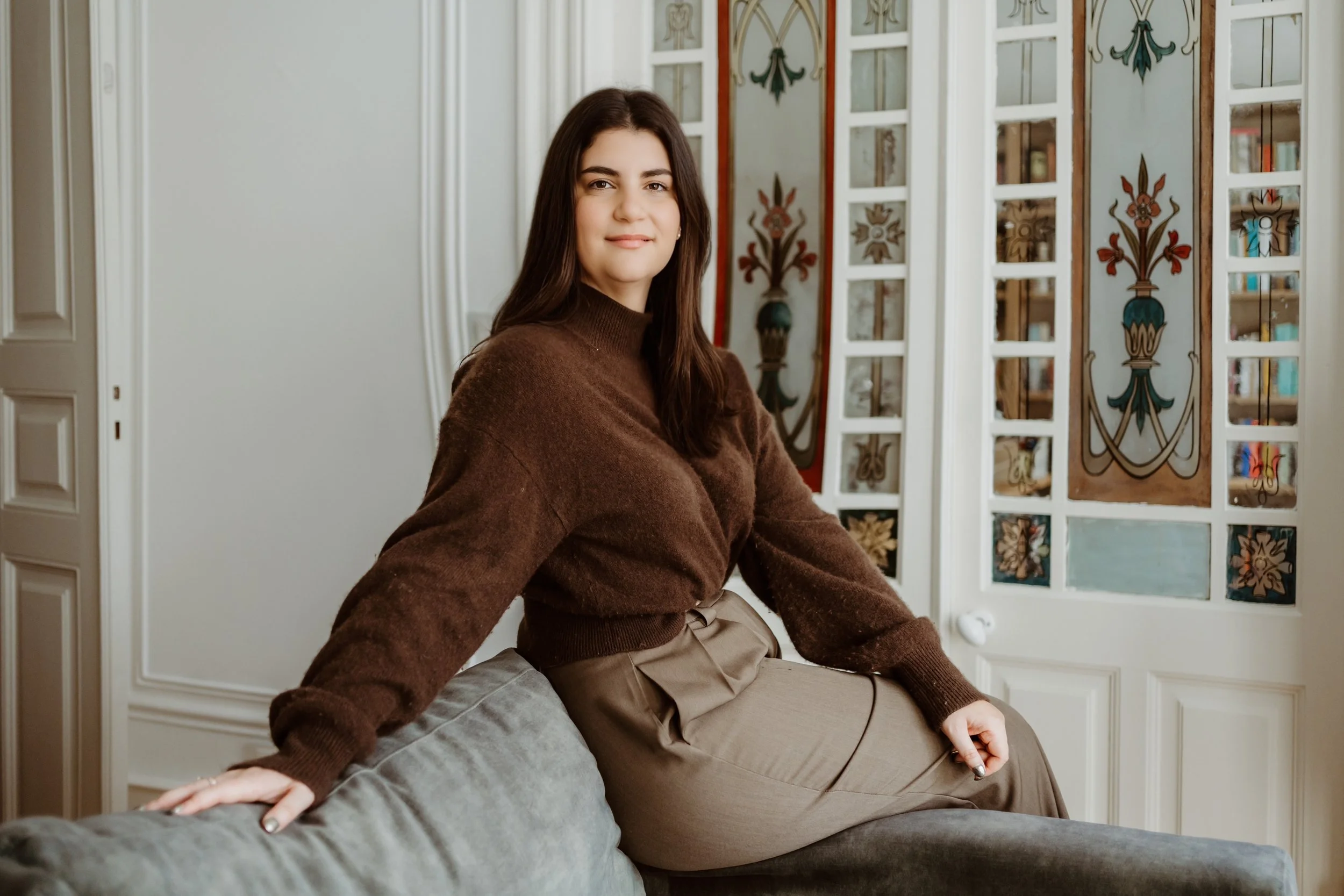 Claudia, founder of Avalon, sitting on the back of a gray sofa, smiling at the camera. She is wearing a brown sweater and beige pants, and is in an office with decorative glass doors with floral designs.