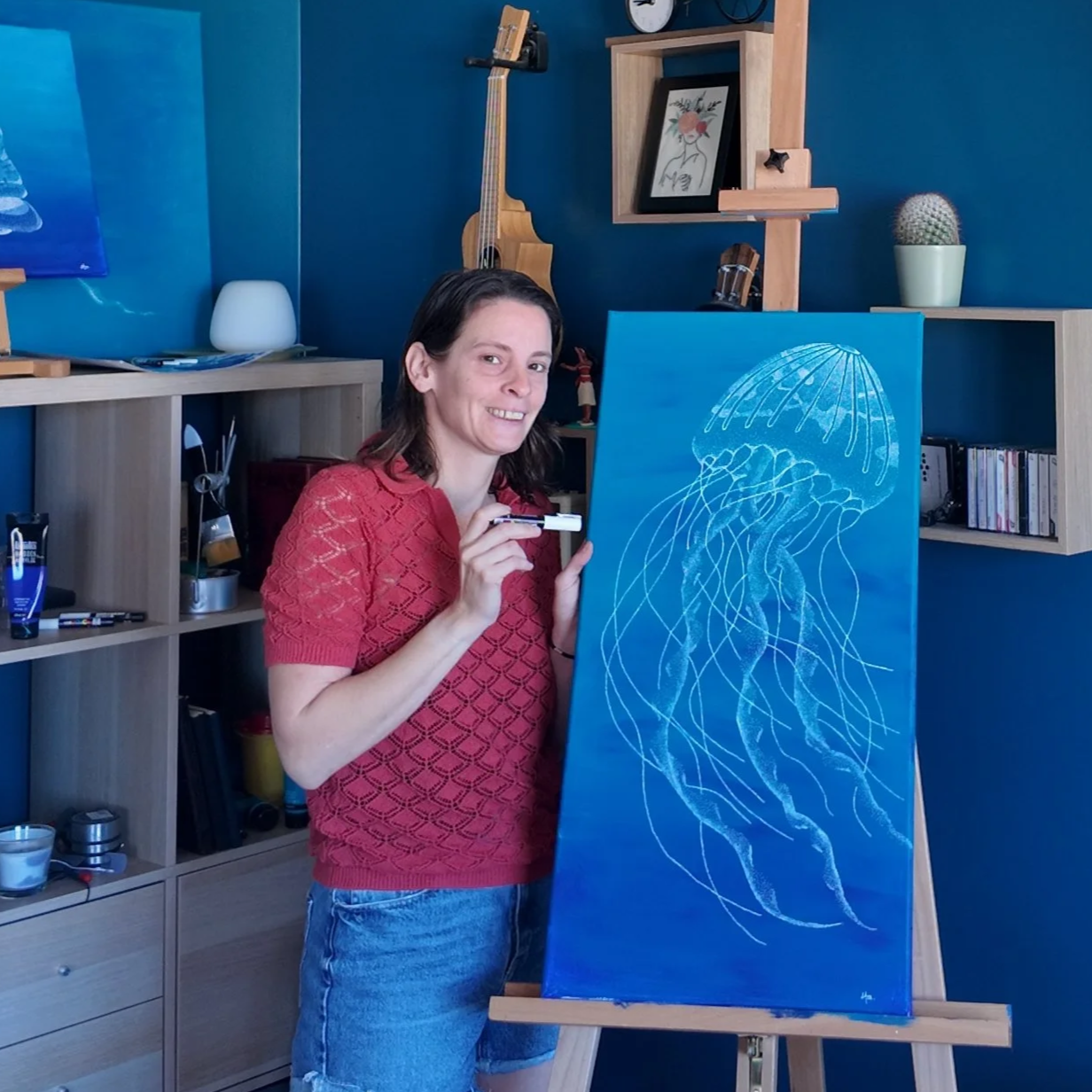 Une femme souriante en train de peindre une toile avec une image de méduse en blanc sur fond bleu dans un intérieur moderne.