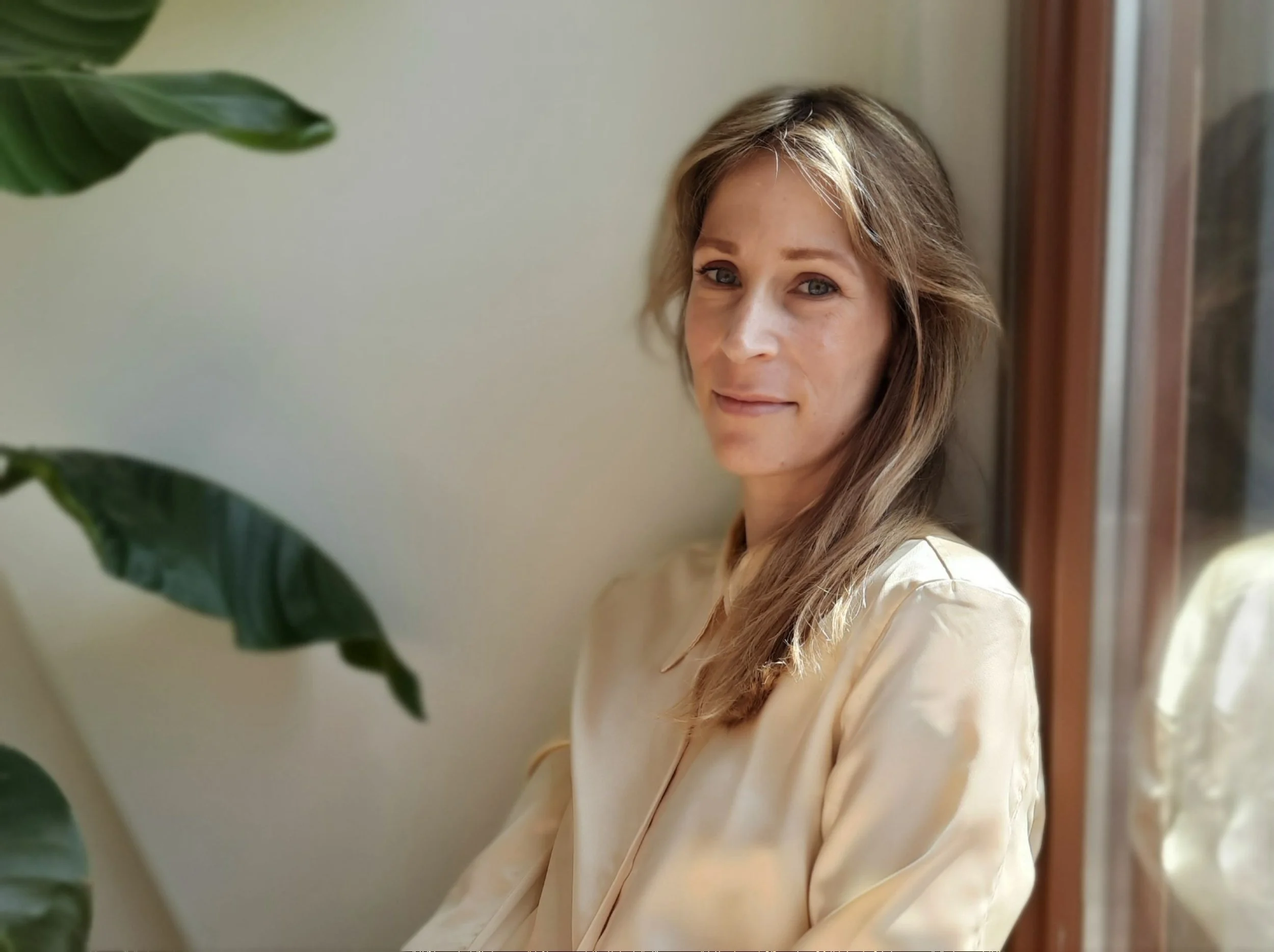 Een vrouw met bruin haar die voor een raam zit, met plantblad zichtbaar in de linkerbovenhoek.