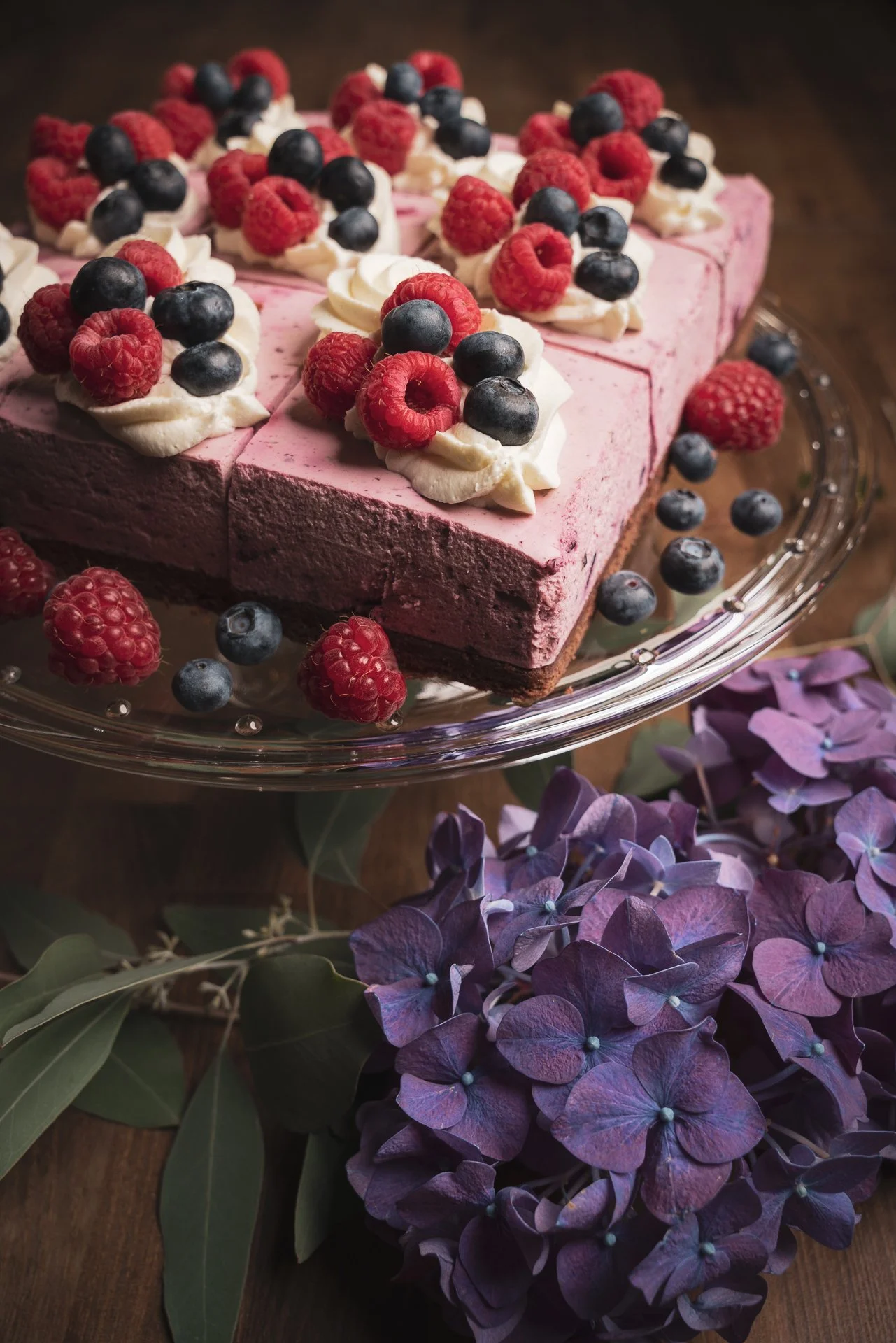 Aki Suomela valokuvaus portfolio. Herkullinen kerrosjuustokakku marjoilla ja kermavaahdolla tarjoilulautasella. Taustalla lilahödyn kukka ja vihreitä lehtiä.