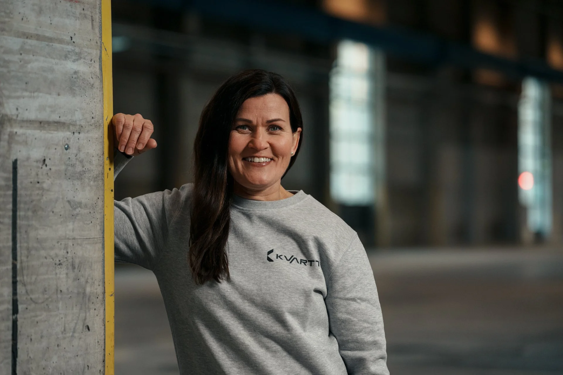 Aki Suomela valokuvaus portfolio. Nainen hymyilee teollisuusalueella, nojaten seinään. Hänellä on harmaa pitkähihainen paita.