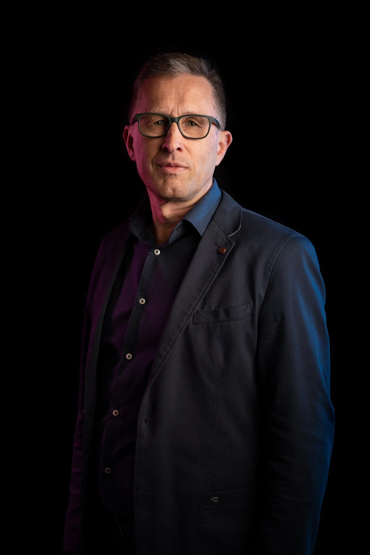 Aki Suomela valokuvaus portfolio. Kuvassa mies puvussa ja silmälaseissa, mustalla taustalla. Hän katsoo suoraan kameraan.