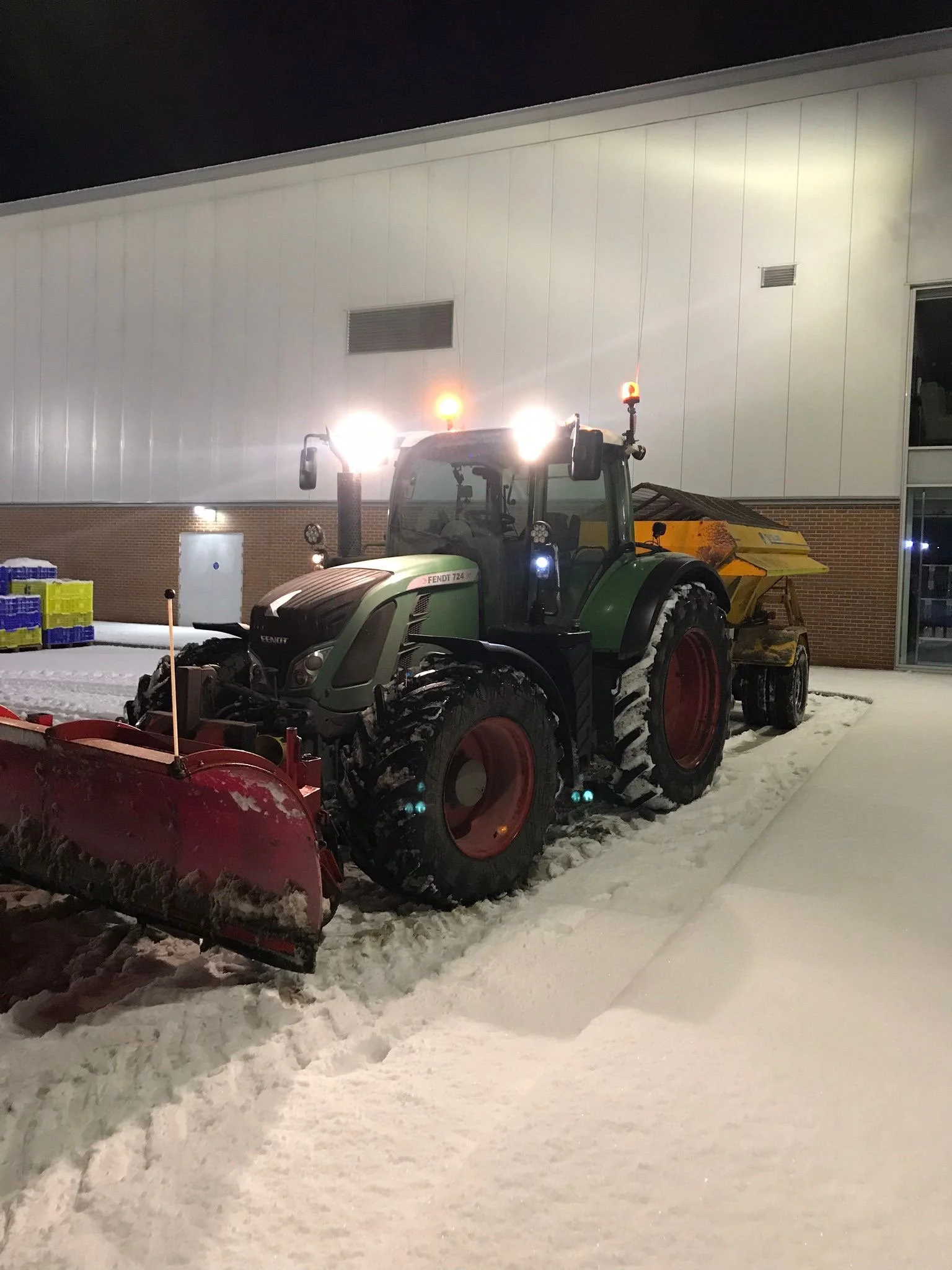 A green tractor equipped with a snow plow attachment clearing snow outside a building at night.