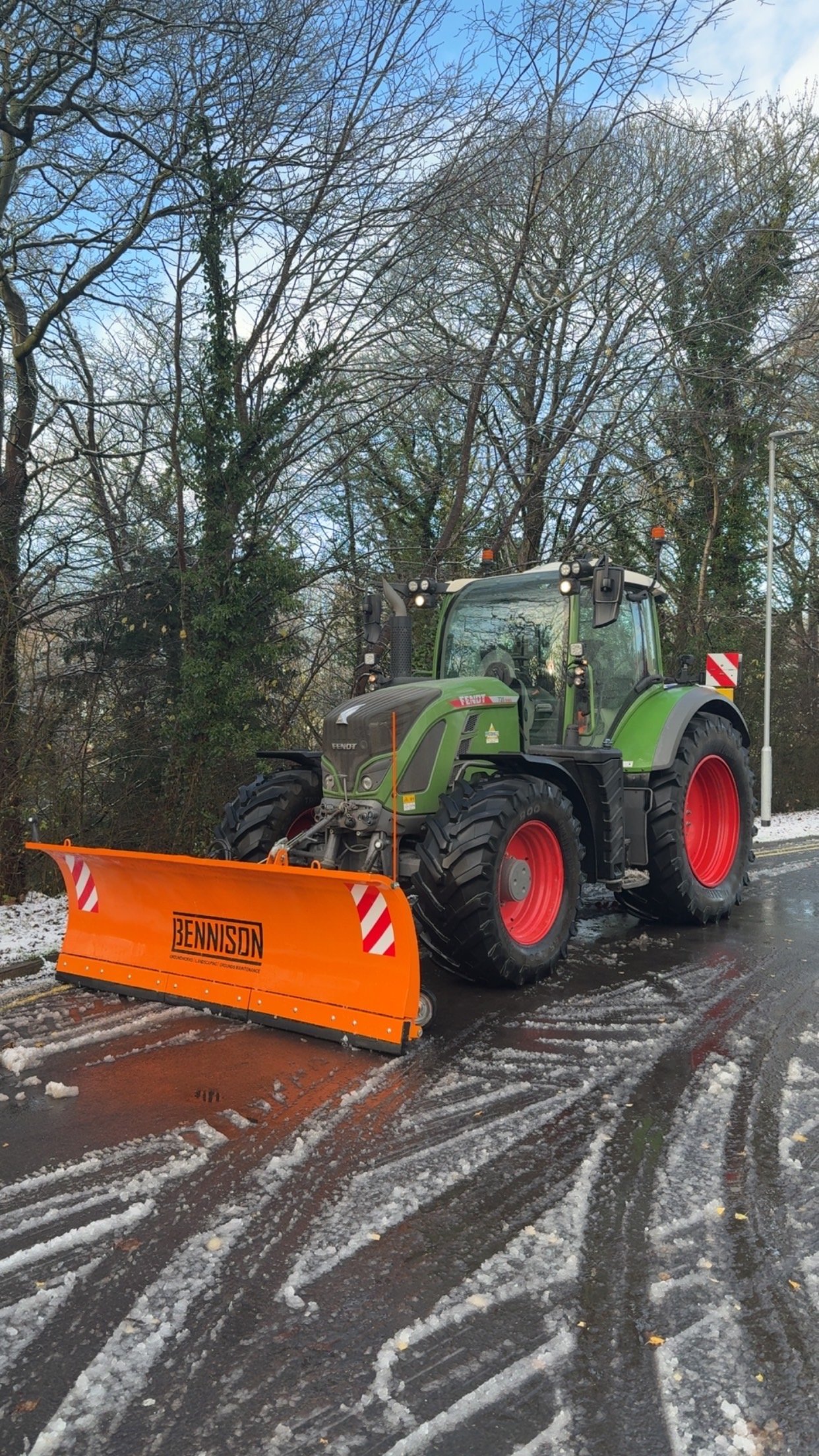A green tractor with red wheels is clearing snow from a road, with an orange snow plow attached at the front, and trees in the background.