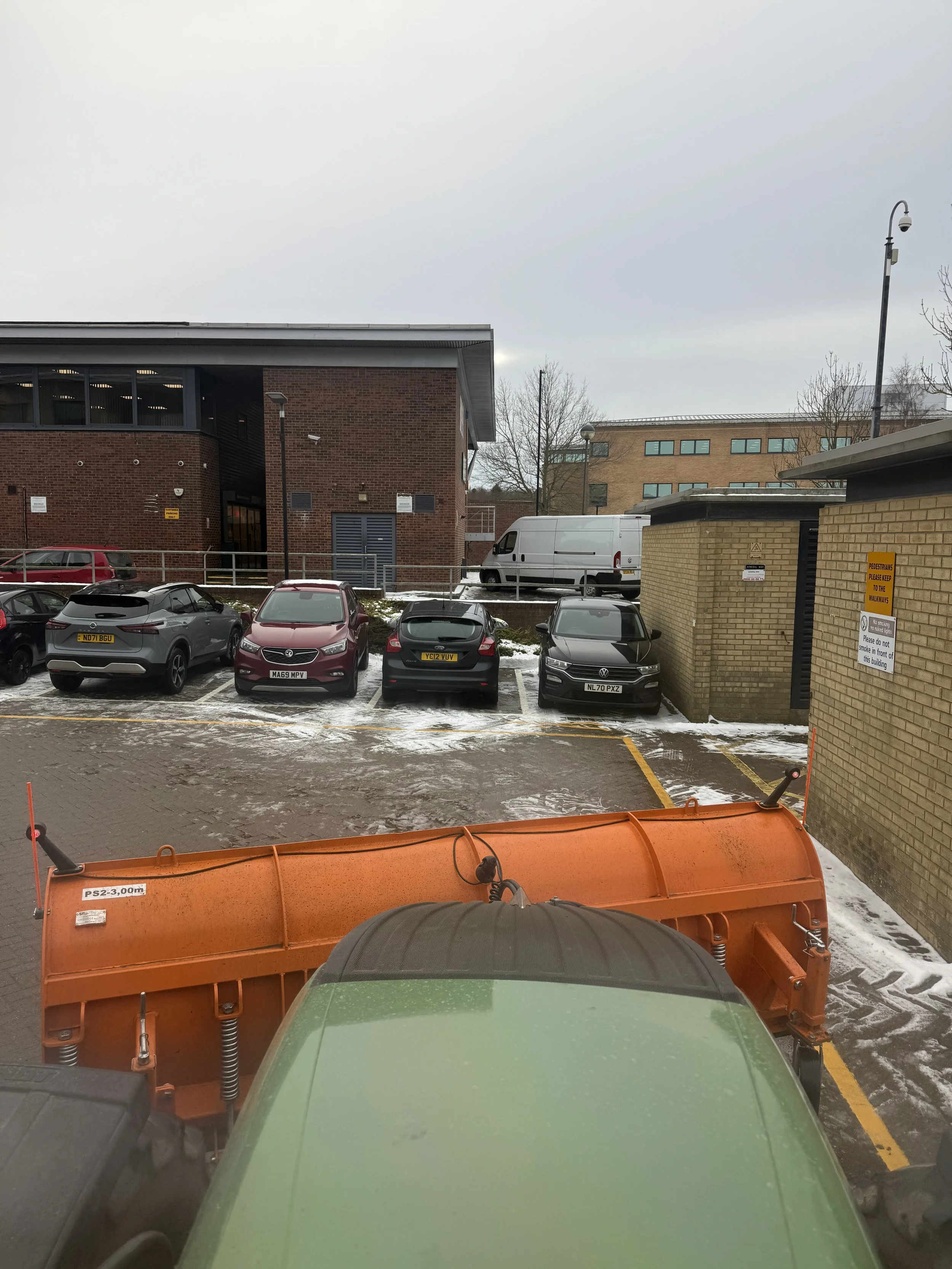 View from inside a vehicle showing a snow-dusted parking lot with several parked cars, a building with brick and glass exterior, a white van, and a snowplow attachment in the foreground.