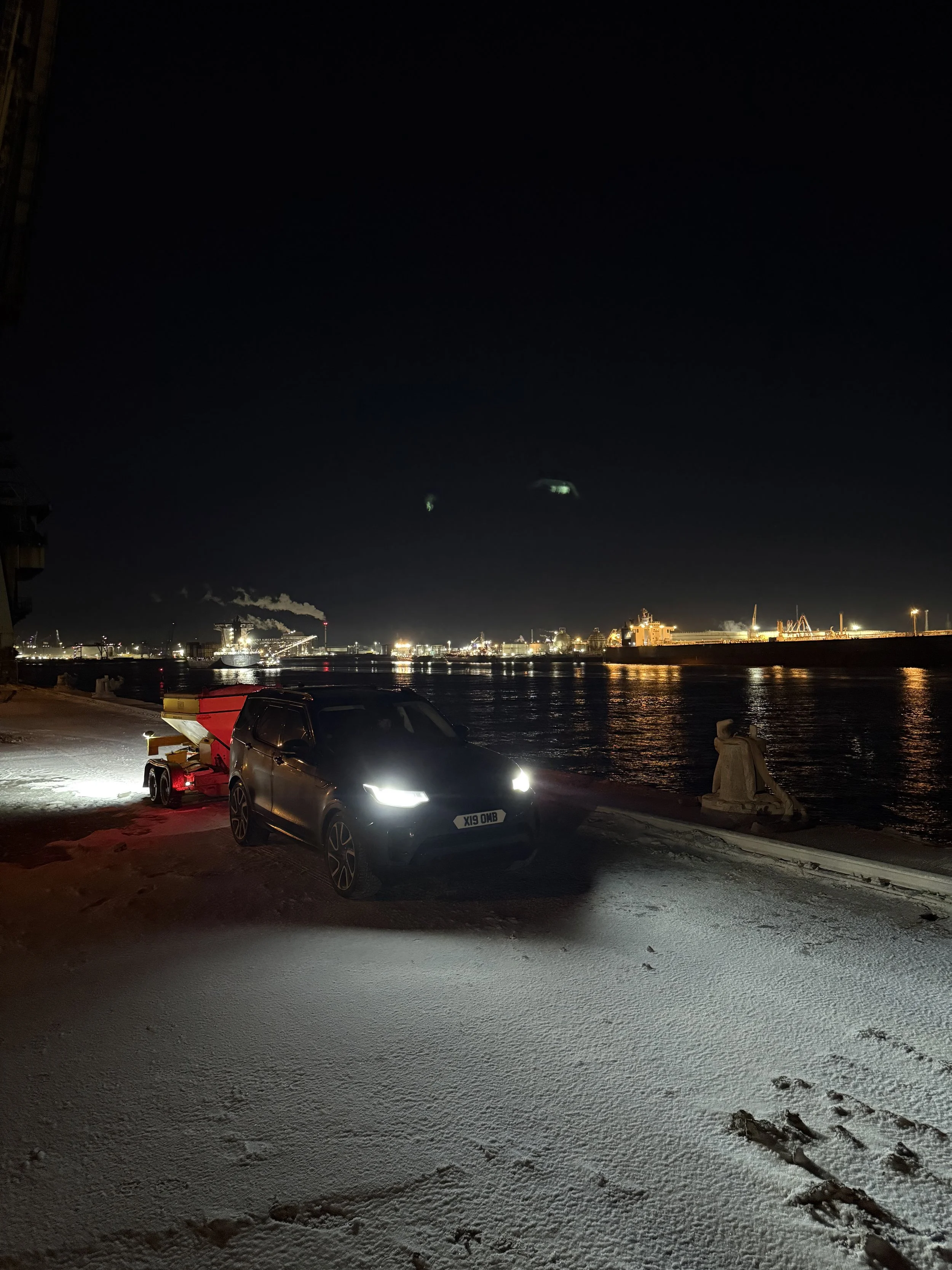 Nighttime scene by the water with a black vehicle and Gritter on snow-covered ground, industrial cityscape with bright lights and smoke in the distance.