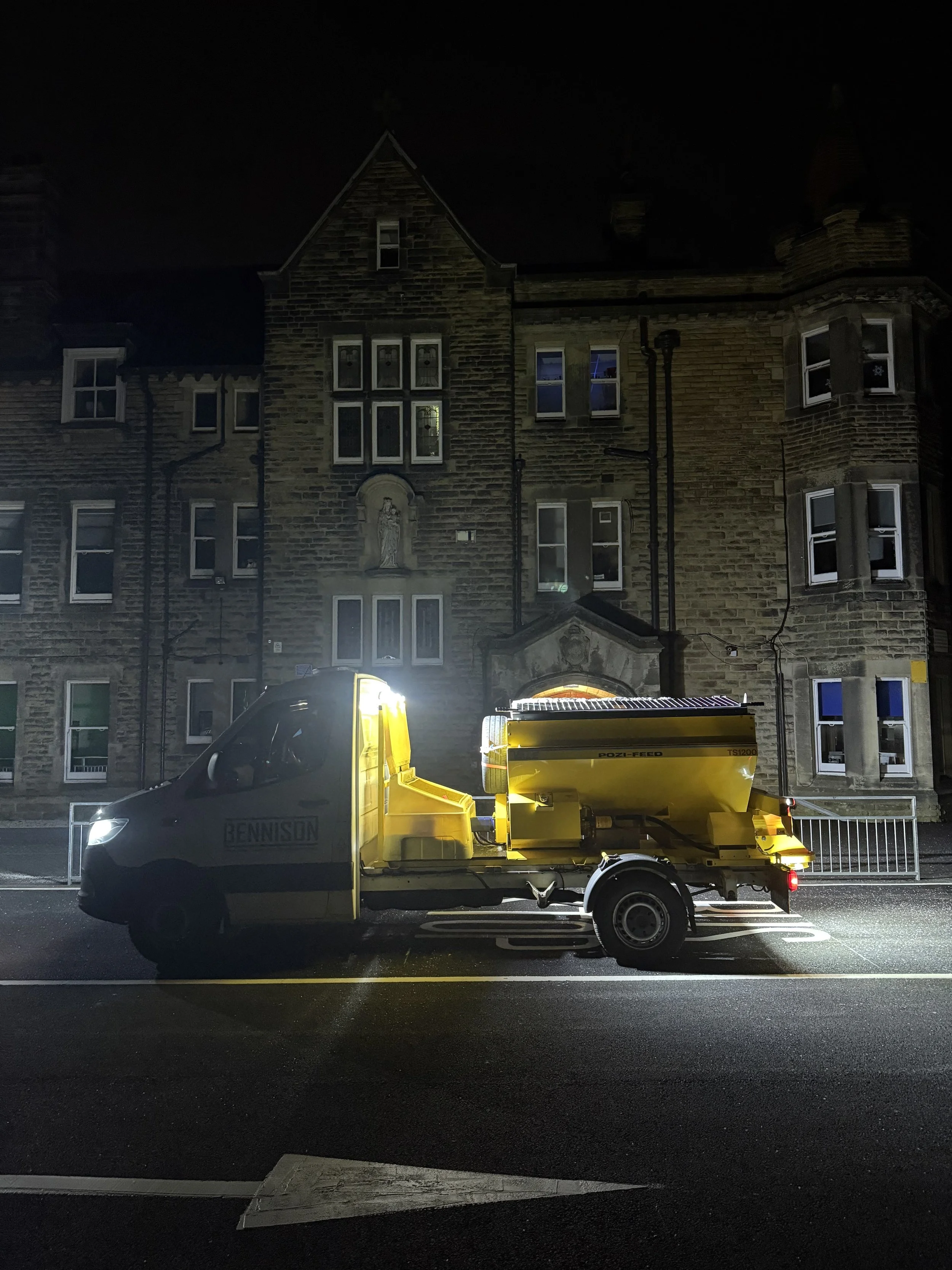 A yellow street cleaning truck parked on a city street at night, with a large historic brick building in the background.