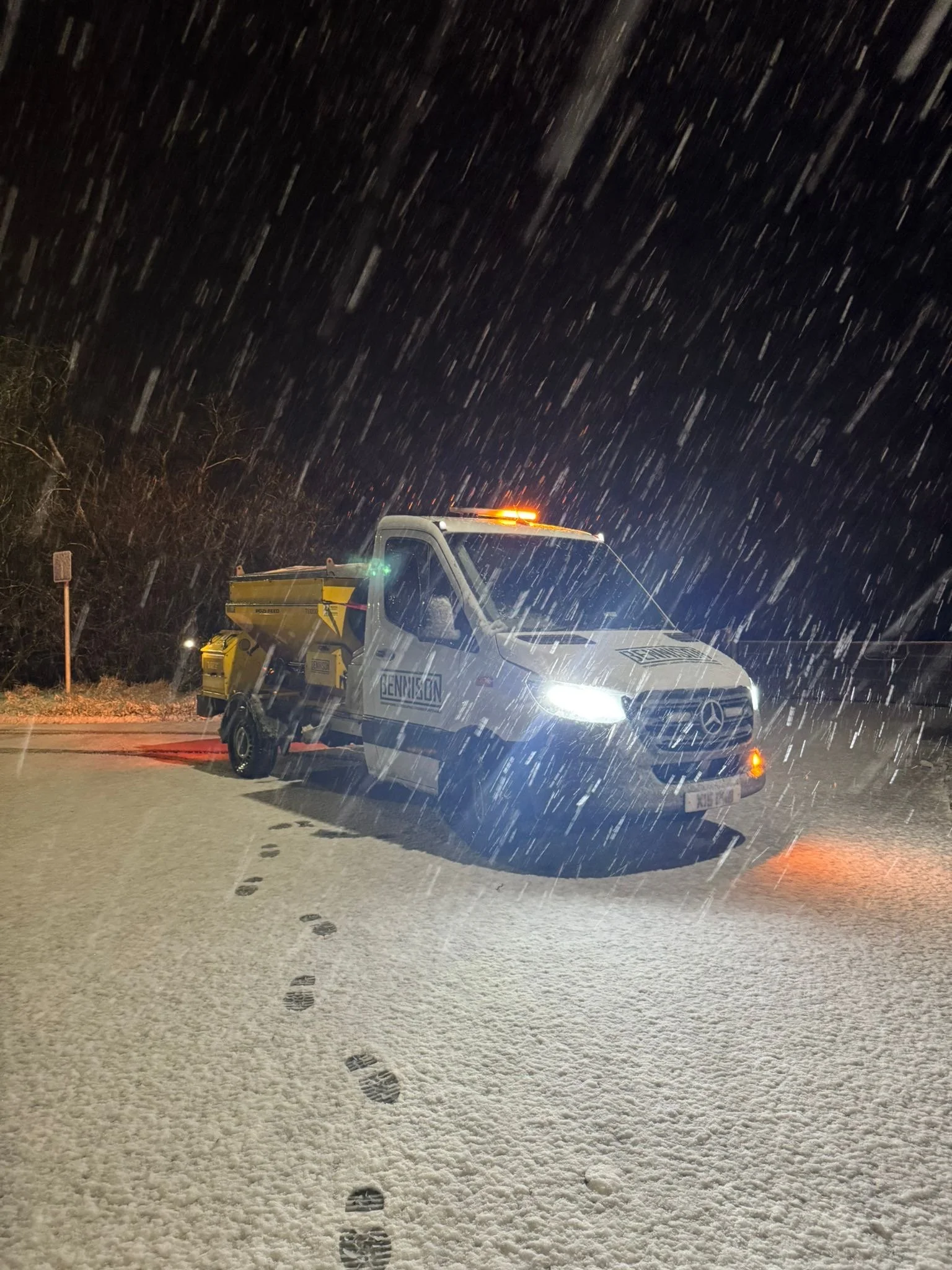 A white truck with a Gritter on the back, side labeled 'BENNISION,' parked on a snow-covered road at night in heavy snowfall, with the truck's headlights on. Ready for gritting car parks