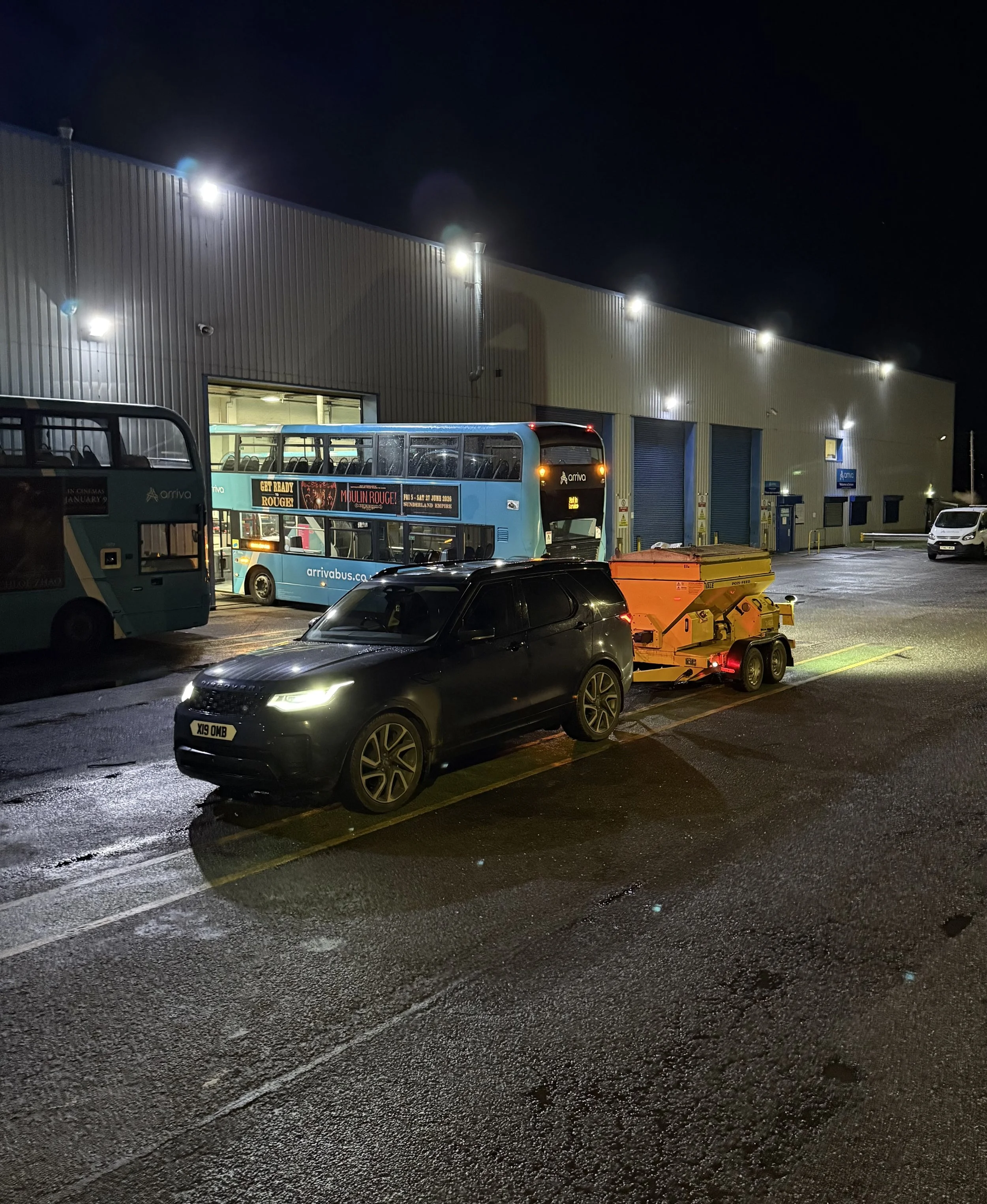 Night scene at a bus depot with a black SUV towing a yellow trailer, two double-decker buses, a white van, and a warehouse with bright exterior lights.