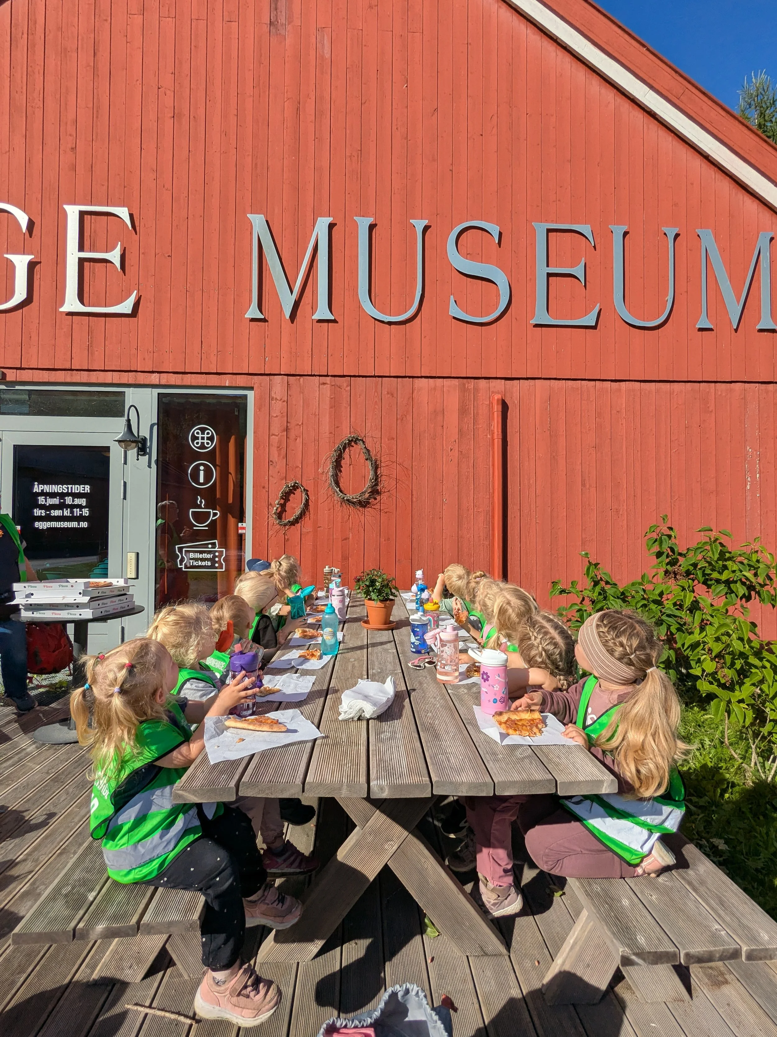 Barn som sitter ved en utendørs benk og spiser mat, foran en gammel rød bygning med skilt som sier 'MUSEUM'.