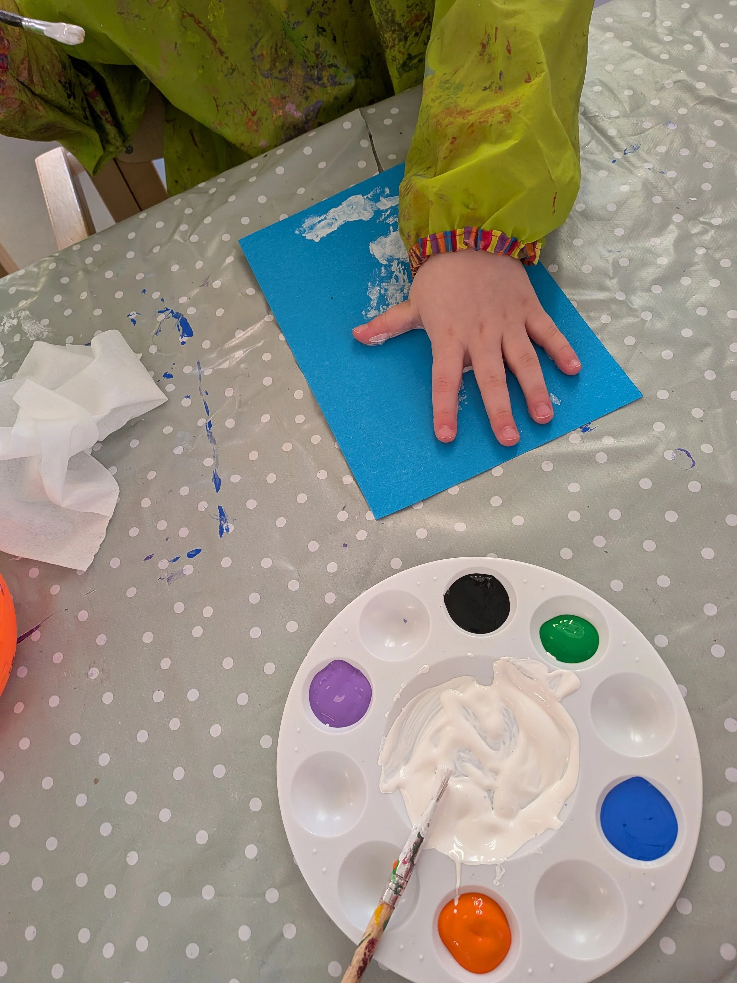 Bilde av en barnehånd som holder i et blått papir. På bordet er en palett med maling i ulike farger