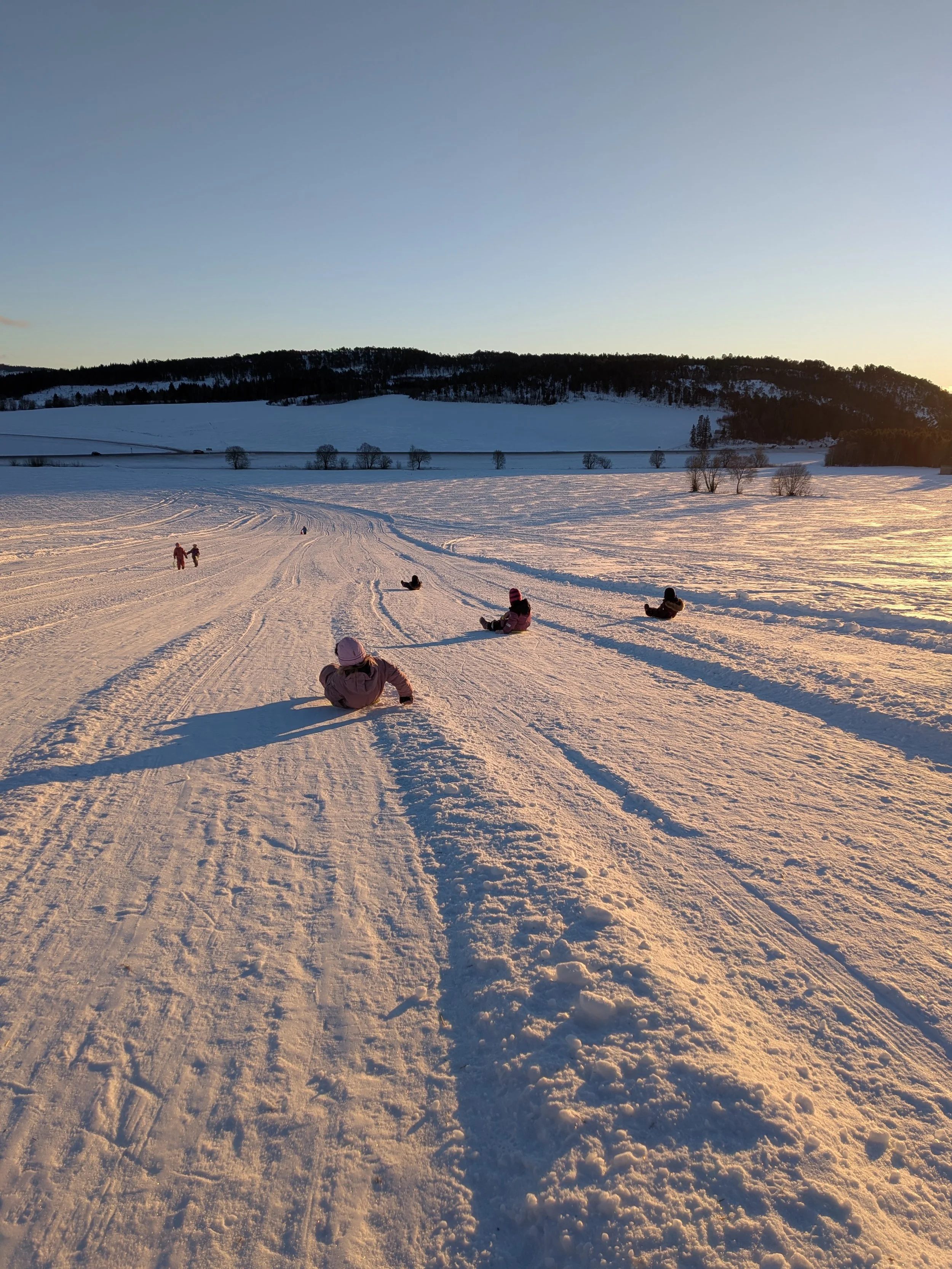 Barn som aker i en snøkledd bakke, med utsikt mot åser, blå himmel og sol.